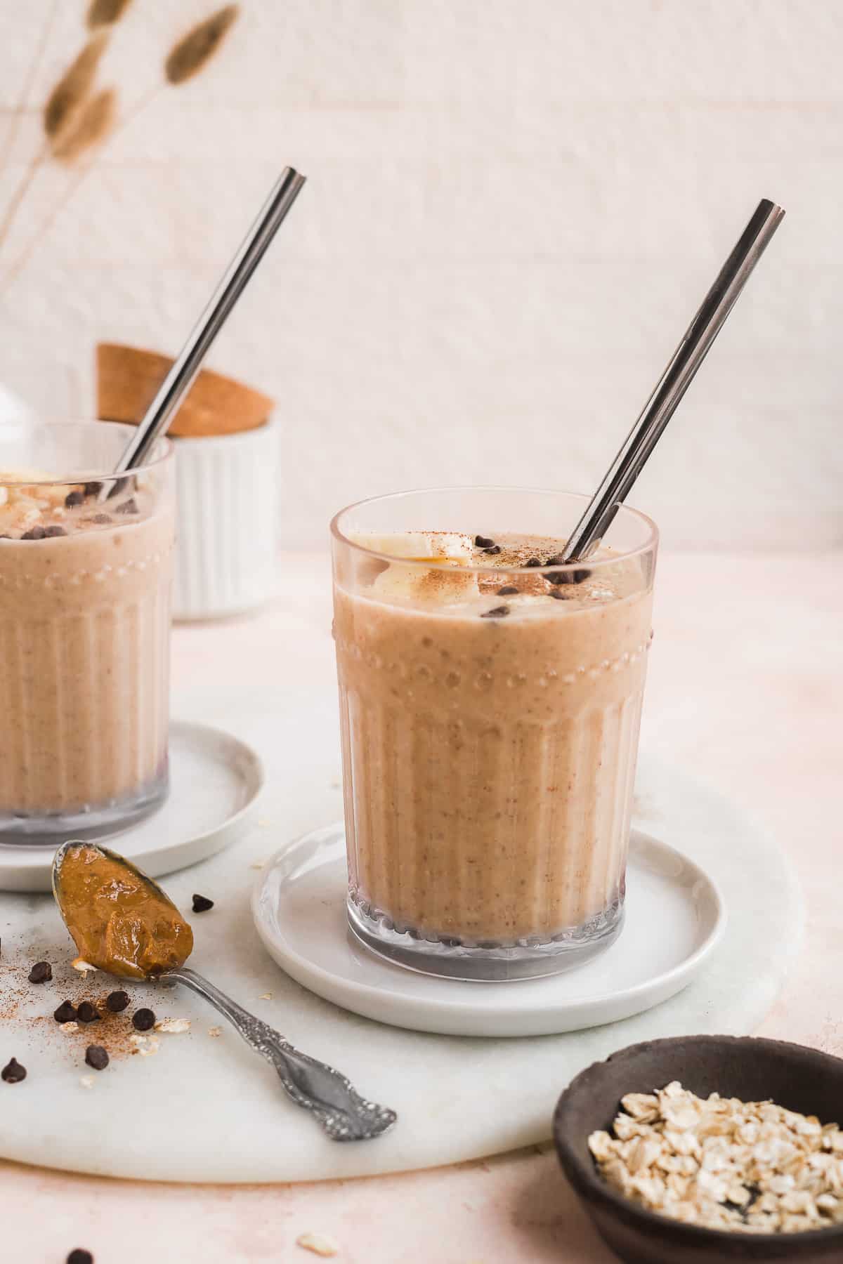 2 clear glasses filled with smoothie with toppings and a straw on white plates with a white background.