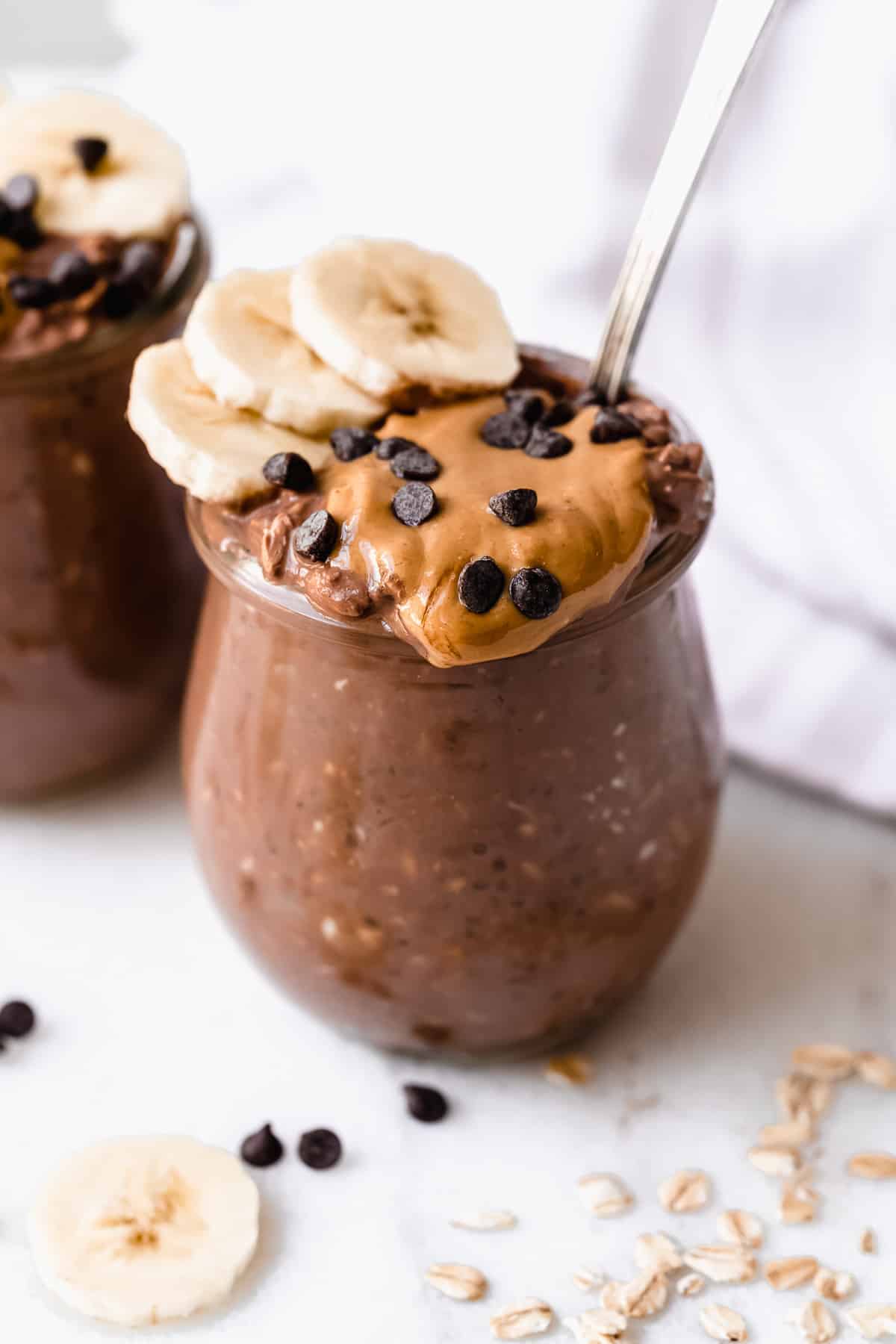 Jar of chocolate oats with peanut butter, banana slices, and chocolate chips on top on a white background