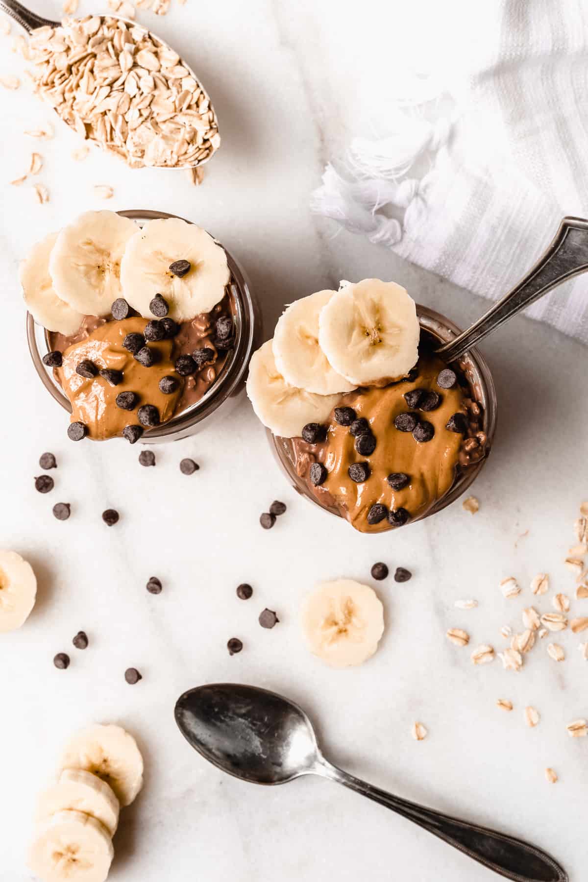 Two jars filled with chocolate oats from above with peanut butter, banana slices, and chocolate chips on top.