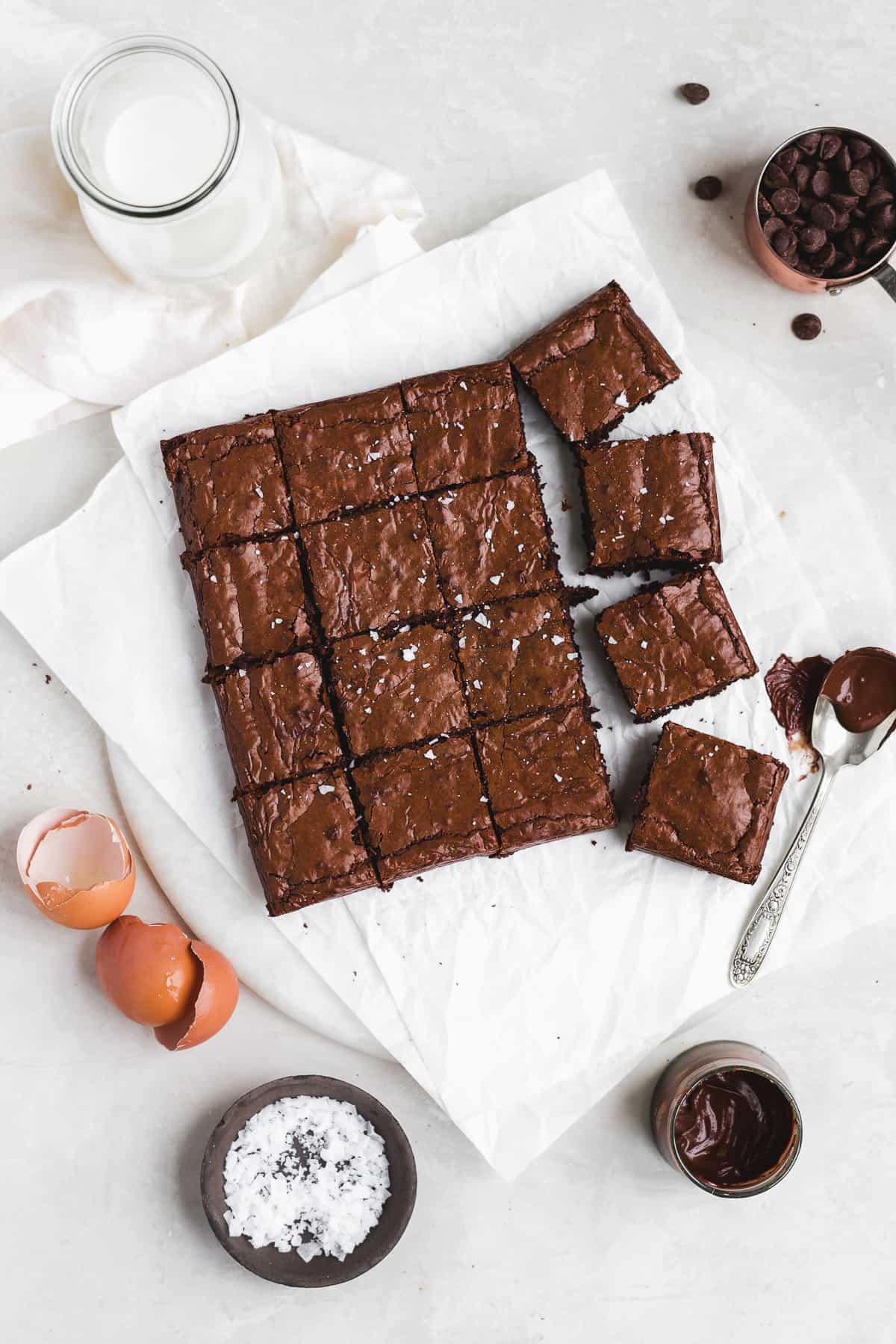 Brownies sliced into squares on a white surface with melted chocolate on the side.