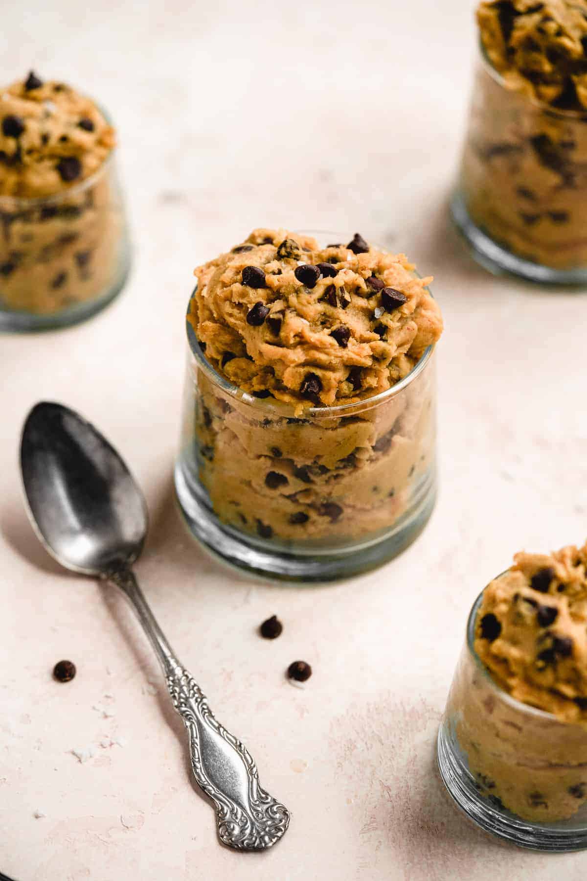 Small clear jar overflowing with yummy edible chocolate chip cookie dough. A silver spoon is resting next to the jar and mini chocolate chips are sprinkled around.