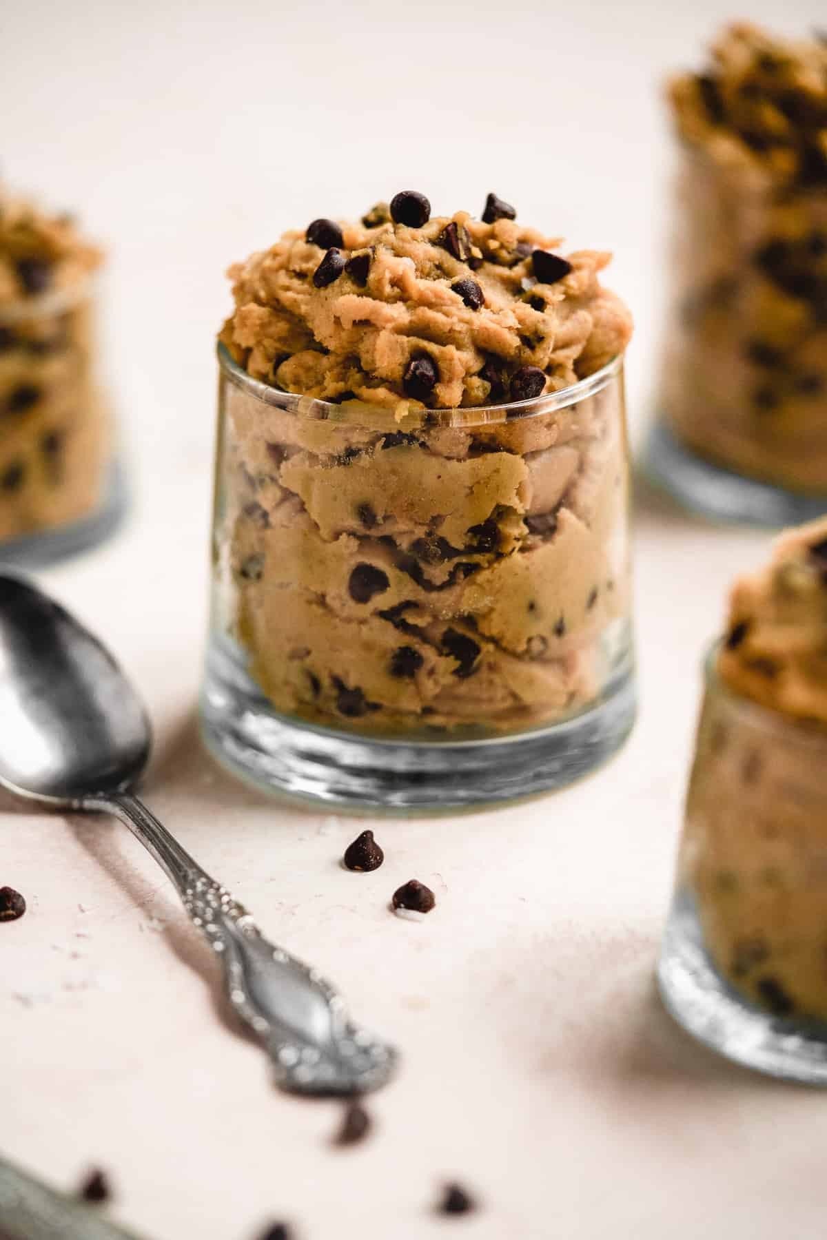 Small clear jar filled to the top with yummy chickpea cookie dough.  Silver spoon is resting beside the jar and chocolate chips are sprinkled around.  