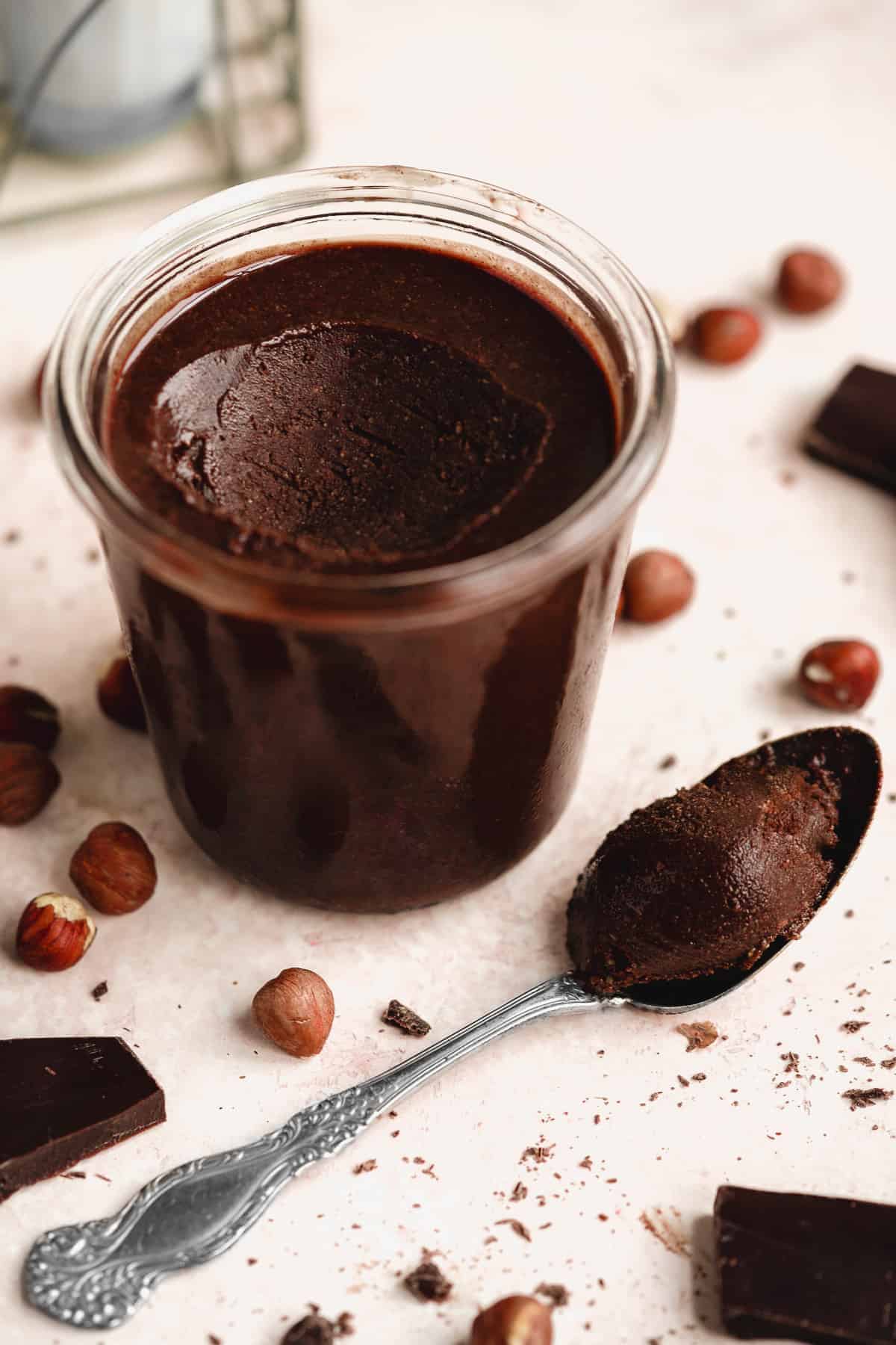 Small clear jar with Chocolate Hazelnut Butter.  A silver spoon with dollop of hazelnut butter is placed beside the jar.  Pieces of chocolate and nuts are sprinkled around.  