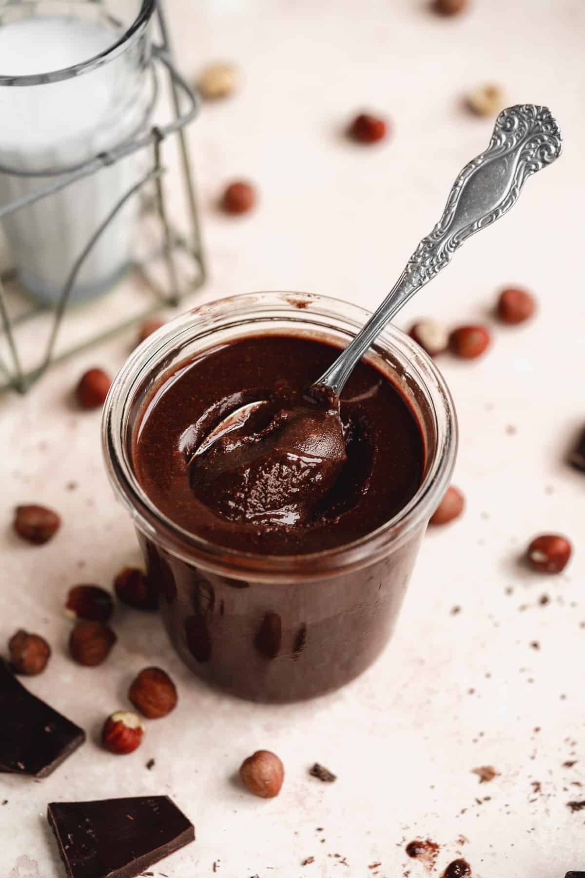 Small jar with delicious hazelnut butter.  Silver spoon dipped inside the jar.  Chocolate pieces and nuts are sprinkled around the jar.  