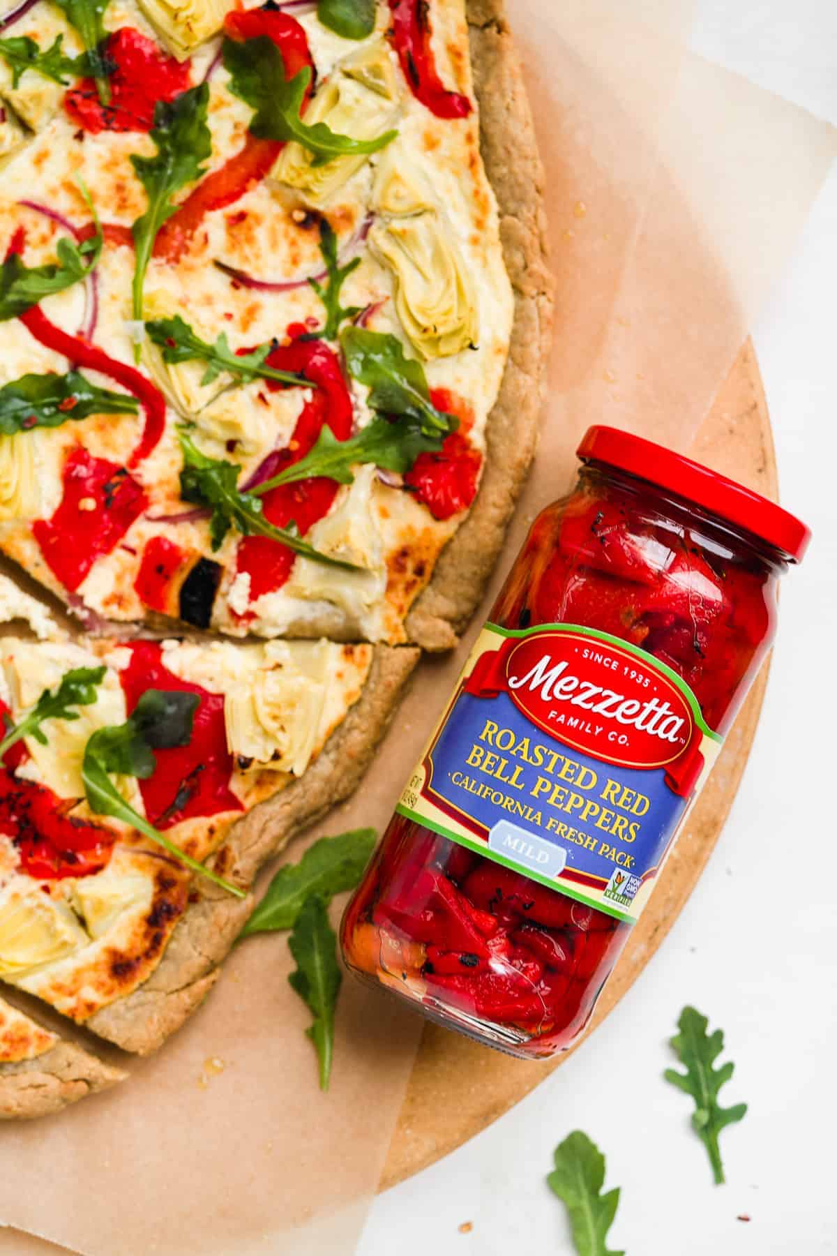 Image of jar of red peppers with a pizza next to it on a brown surface.