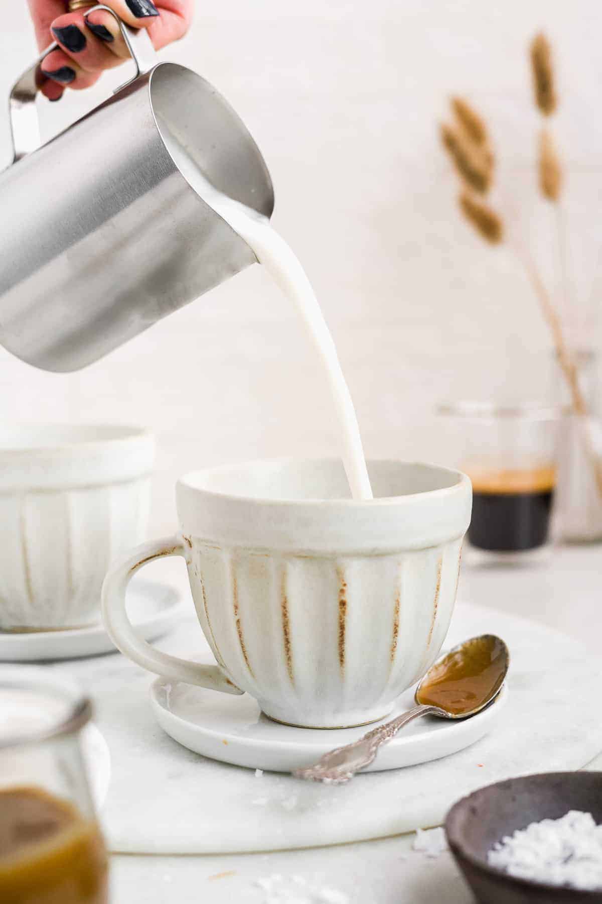 Hand pouring frothed oat milk into a coffee mug on a white surface.