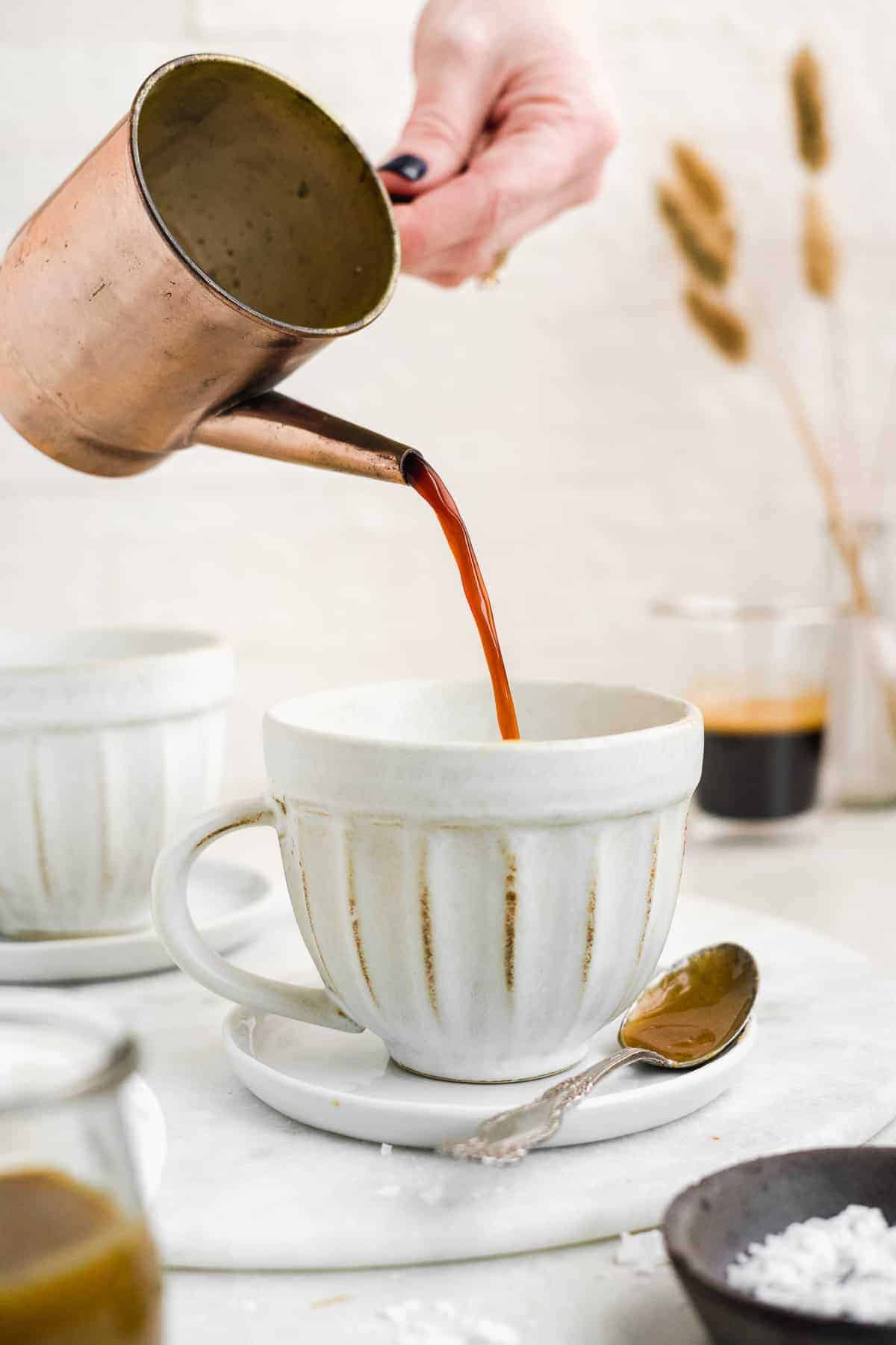 Hand pouring espresso into a coffee mug on a white surface.