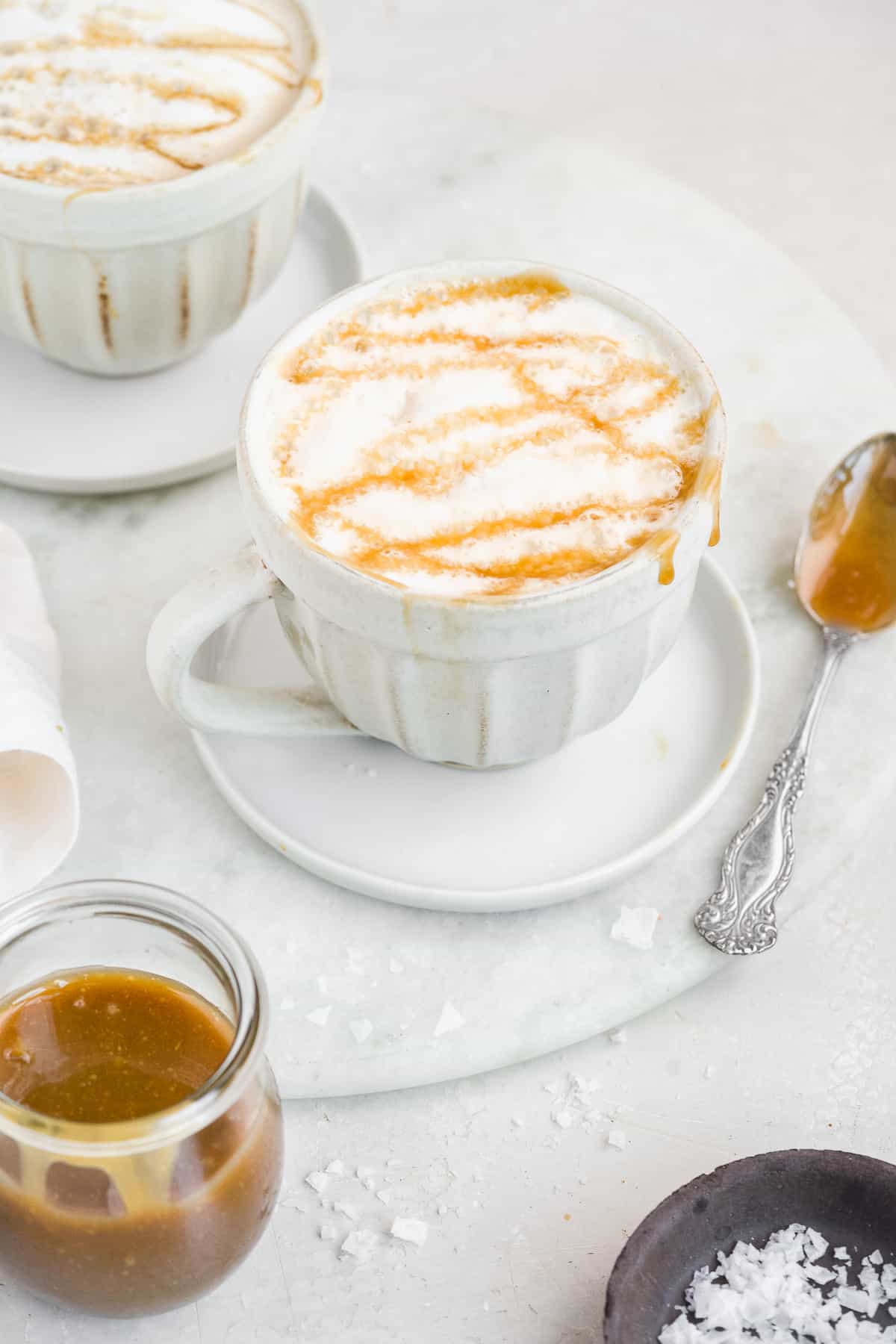 Two white mugs with frothed milk and caramel dripping over the sides on white marble.