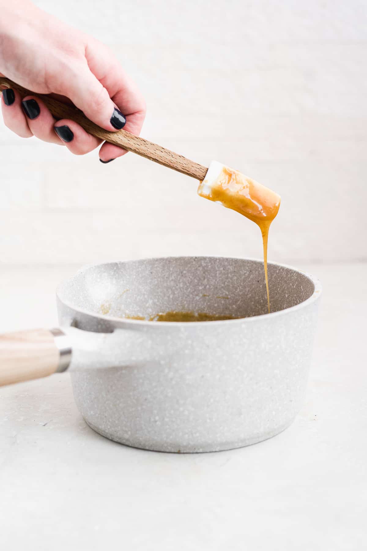 Hand showing caramel dripping off spatula over a white pot.