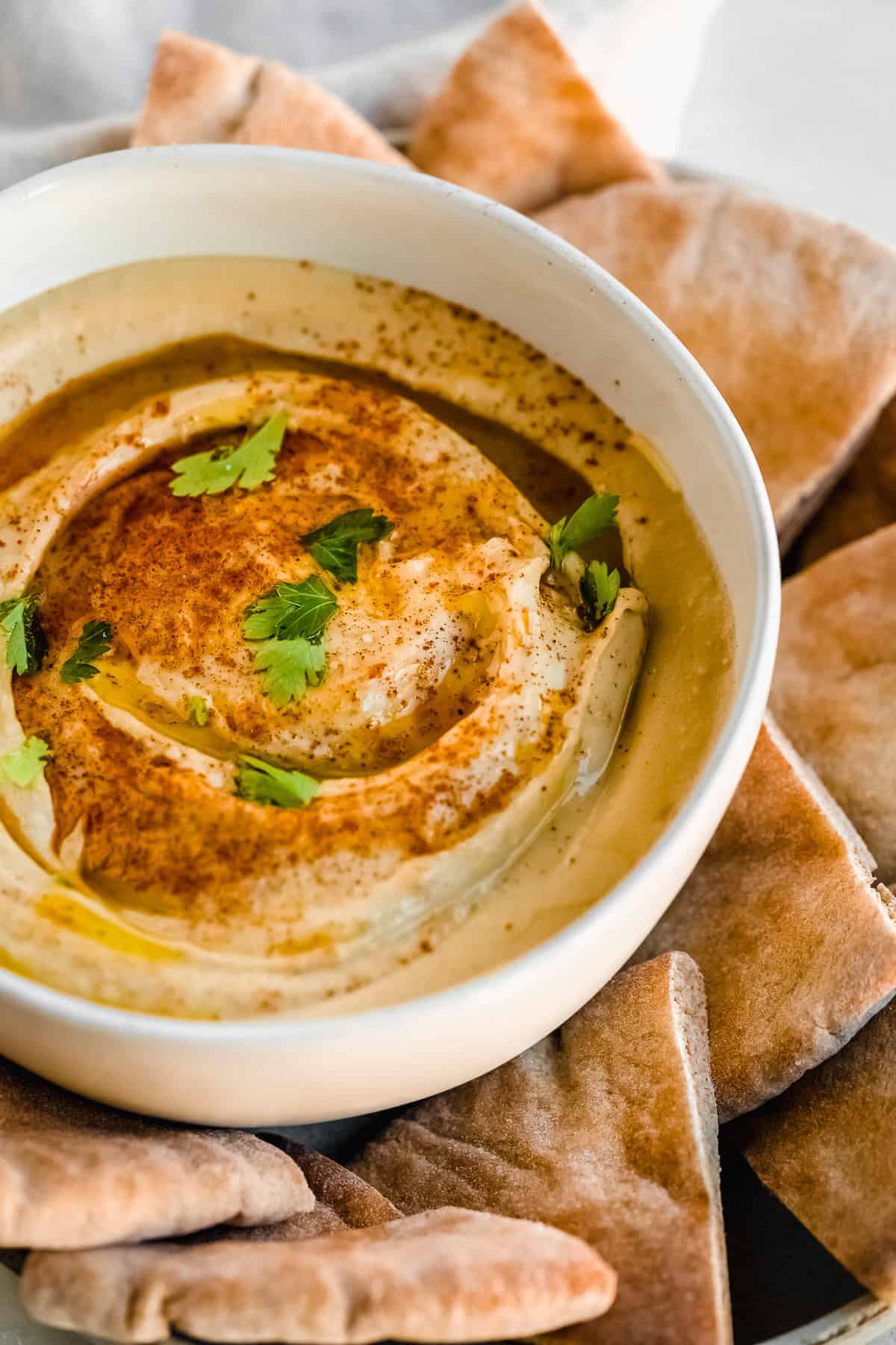 White bowl of hummus topped with green parsley and seasonings on a plate full of pita.