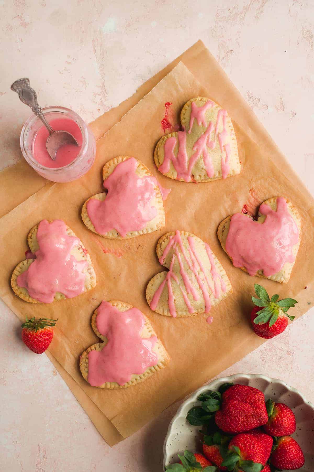 Heart shaped pop tarts on brown parchment paper with pink icing and strawberries in the corner and a jar of pink icing with a spoon.