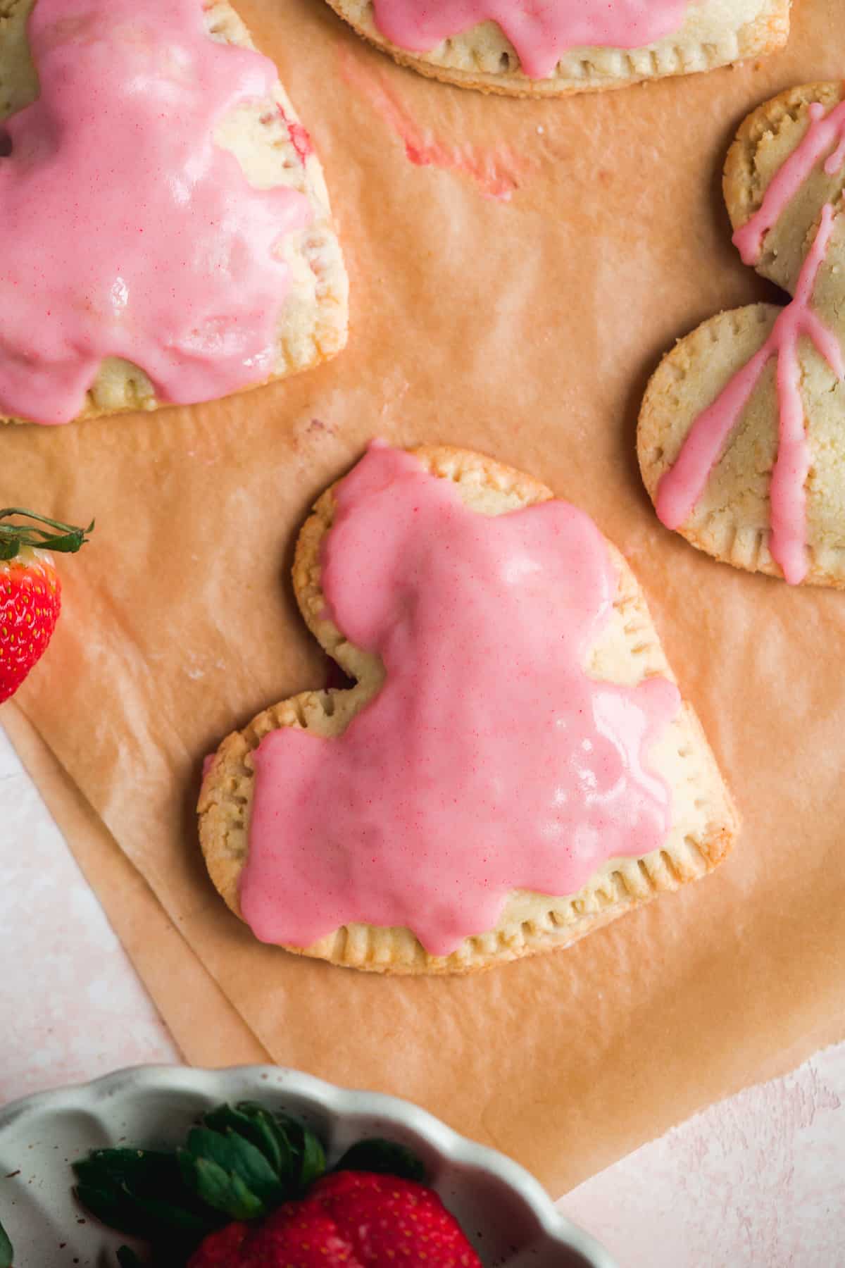 Heart shaped pop tarts on brown parchment paper with pink icing and strawberries in the corner.