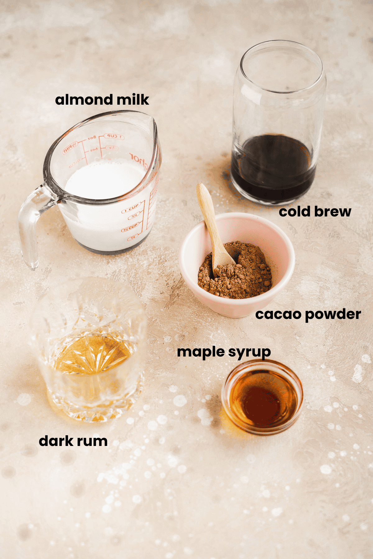 A flat lay of labeled ingredients for a chocolate espresso martini: a glass of cold brew, almond milk in a measuring cup, cacao powder in a pink bowl with spoon, dark rum in a glass, and maple syrup in a small dish.