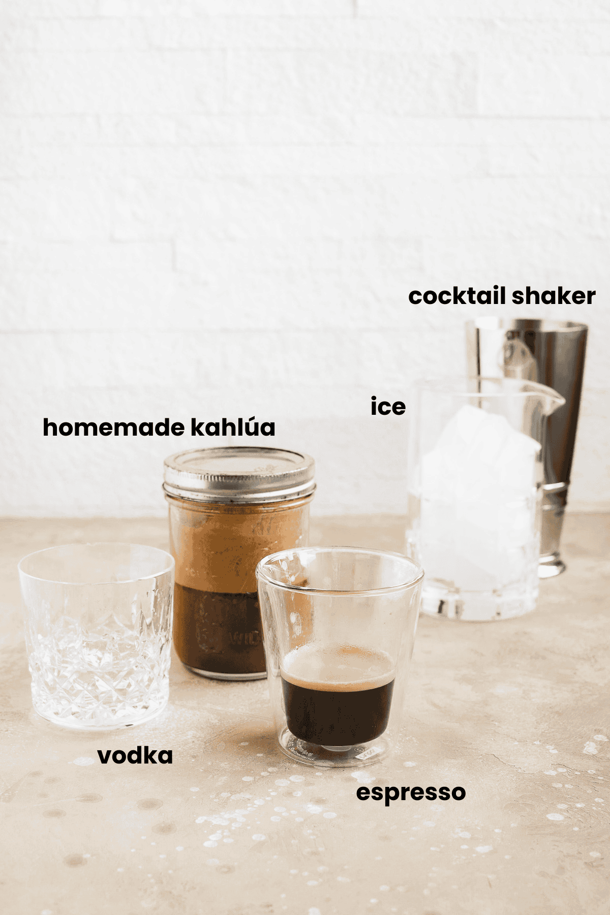 A display of ingredients for a chocolate espresso martini: a glass of vodka, homemade kahlúa in a jar, a glass of espresso, a glass with ice, and a cocktail shaker, each labeled with black text on a light background.