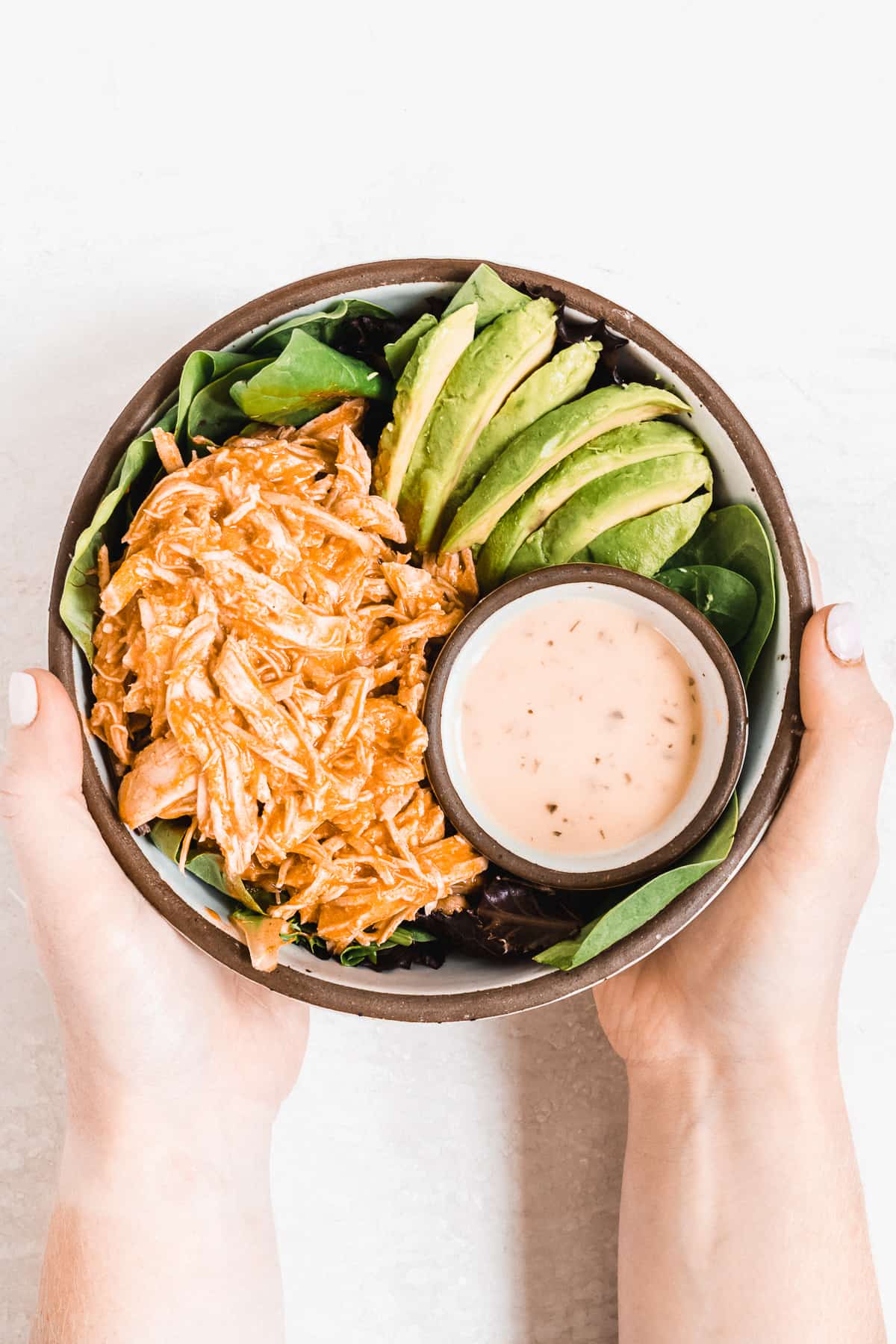 Hands holding bowl full of buffalo chicken, avocado slices, and dressing.