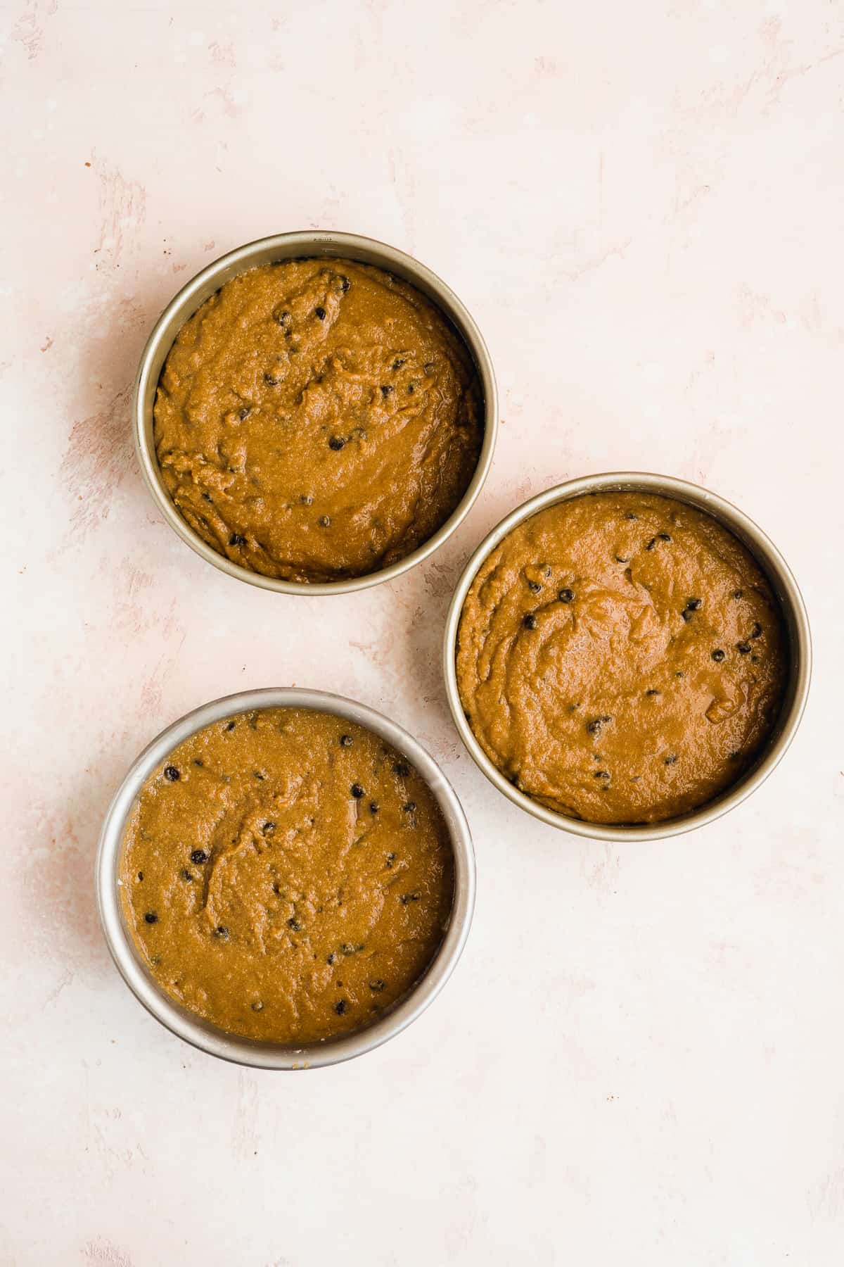 Cookie dough cake batter in three circular cake pans about to be baked.