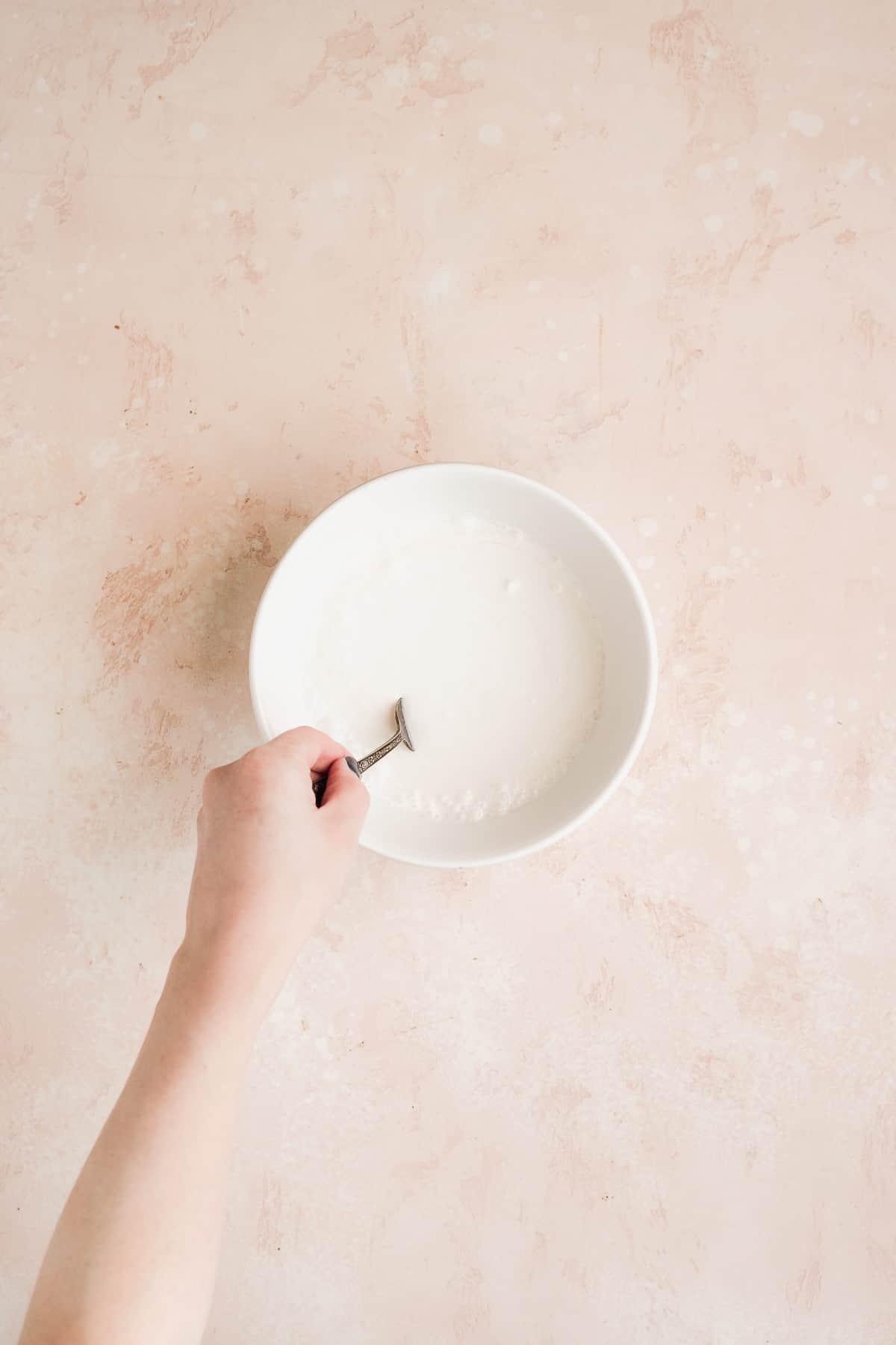 Hand stirring white mixture in a bowl on pink surface.