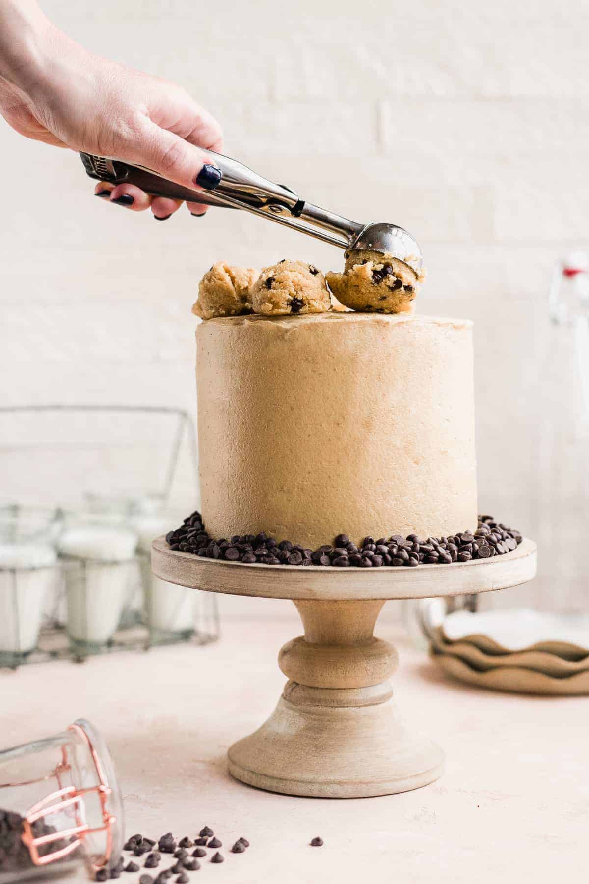 Completed cake sitting on a wooden cake plate with a hand dropping scoop of cookie dough on top. Tiny chocolate chips surround the bottom of the cake. 