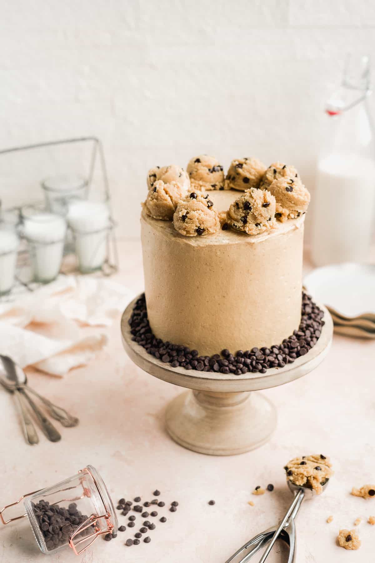 Completed Chocolate Chip Cookie Dough Cake sitting on top of a wooden cake plate with 8 cookie dough scoops on top of the cake. Glasses of milk are in the background.