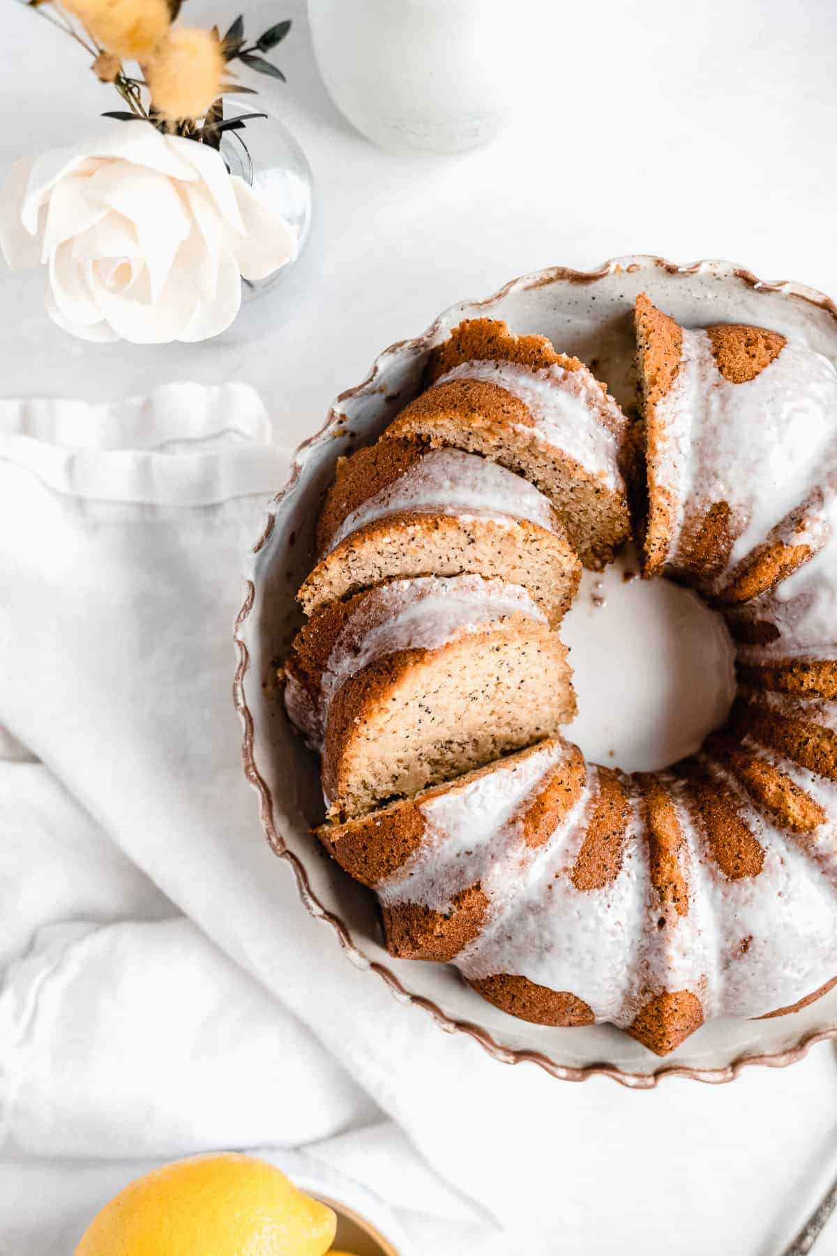 Overhead photo of a freshly baked and iced Citrus-y Gluten Free Lemon Poppy Seed Bundt Cake sitting on a cake plate with several pieces sliced and ready to be enjoyed.  A vase of flowers and a carafe of milk can be seen in the background.  