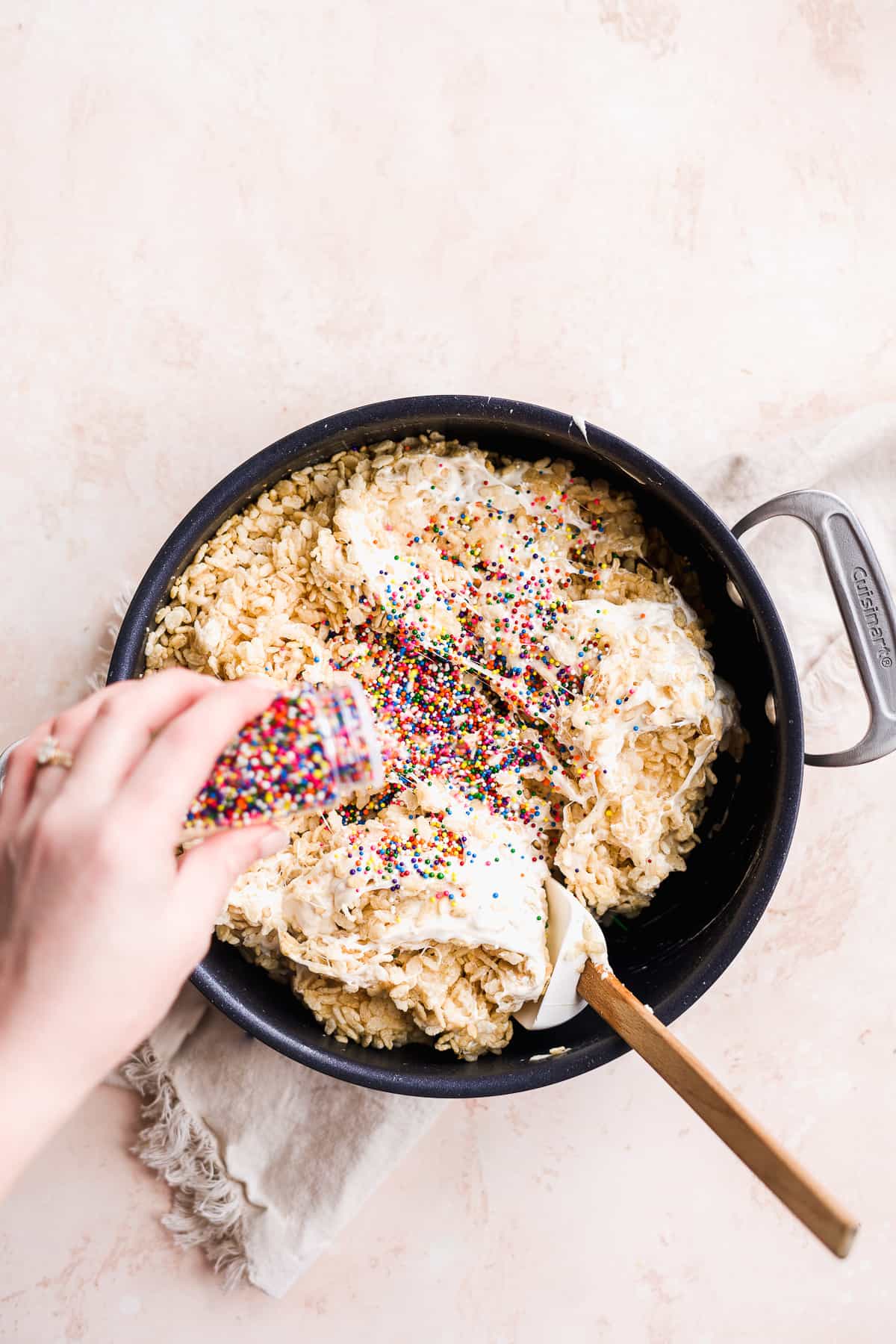 Hand pouring sprinkles into pot of rice krispies and marshmallow fluff.
