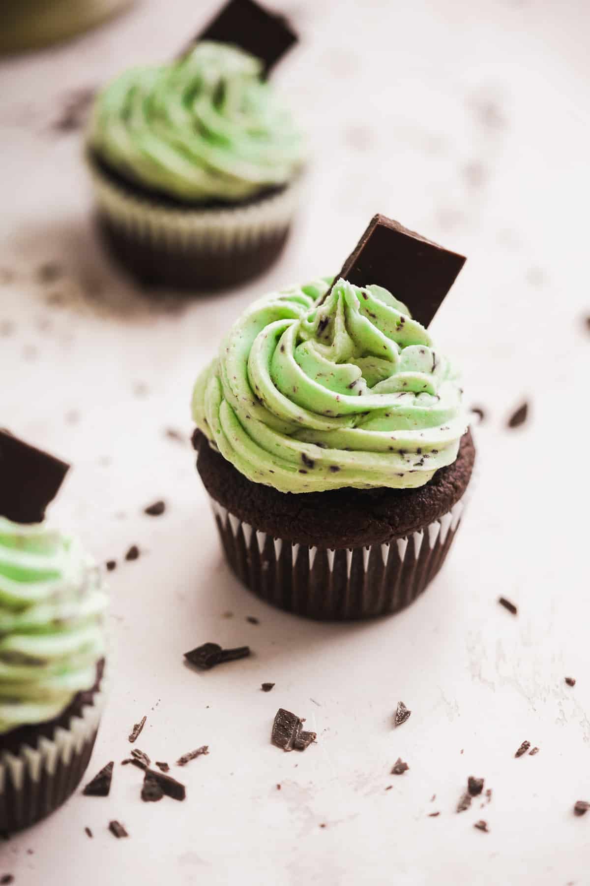 Three chocolate cupcakes with mint green frosting and a chocolate square on top sitting on a cream surface.