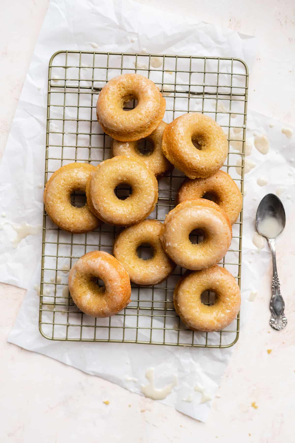 Glazed donuts piled on a wire rack with white paper underneath and a spoon on the side.