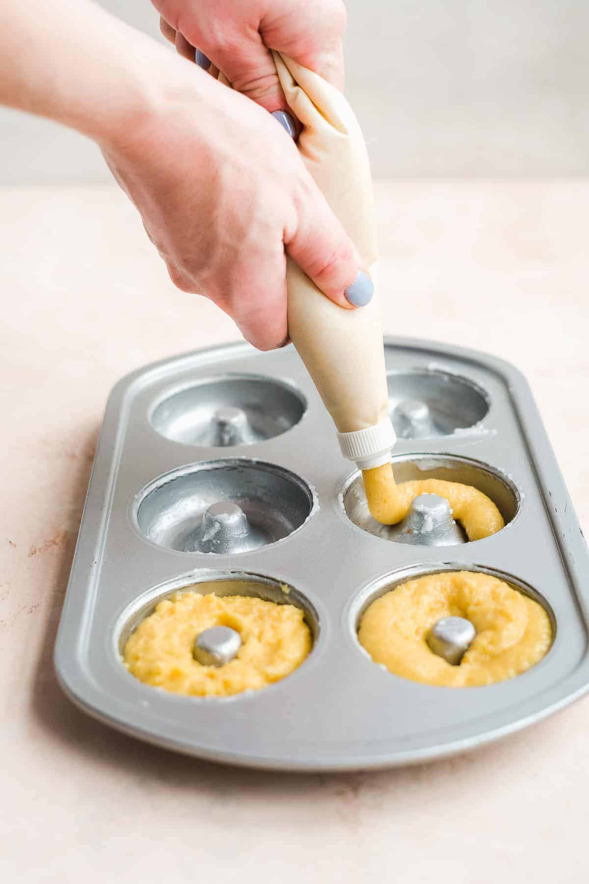 Woman pipping batter into donut mold on a pink surface.