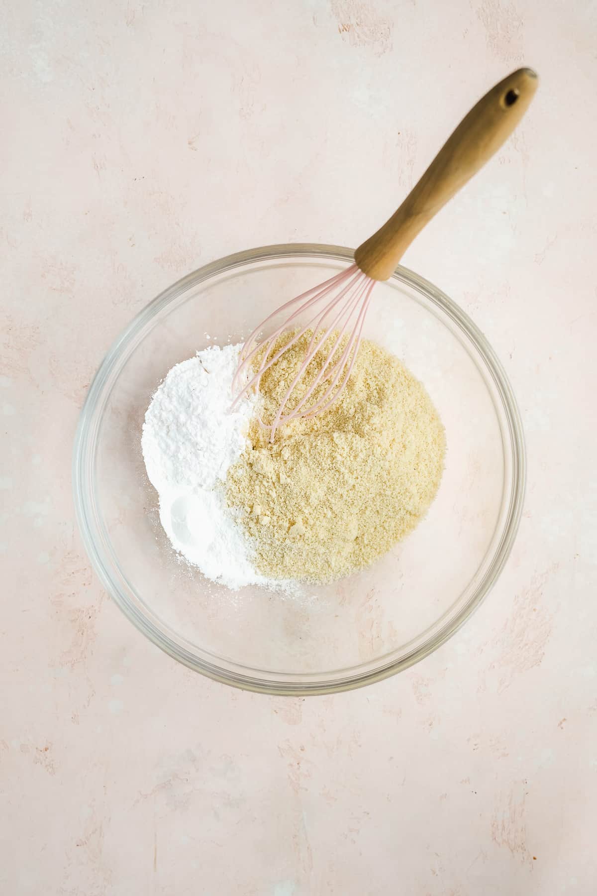 Glass bowl with flour ingredients and a whisk on a light pink surface.