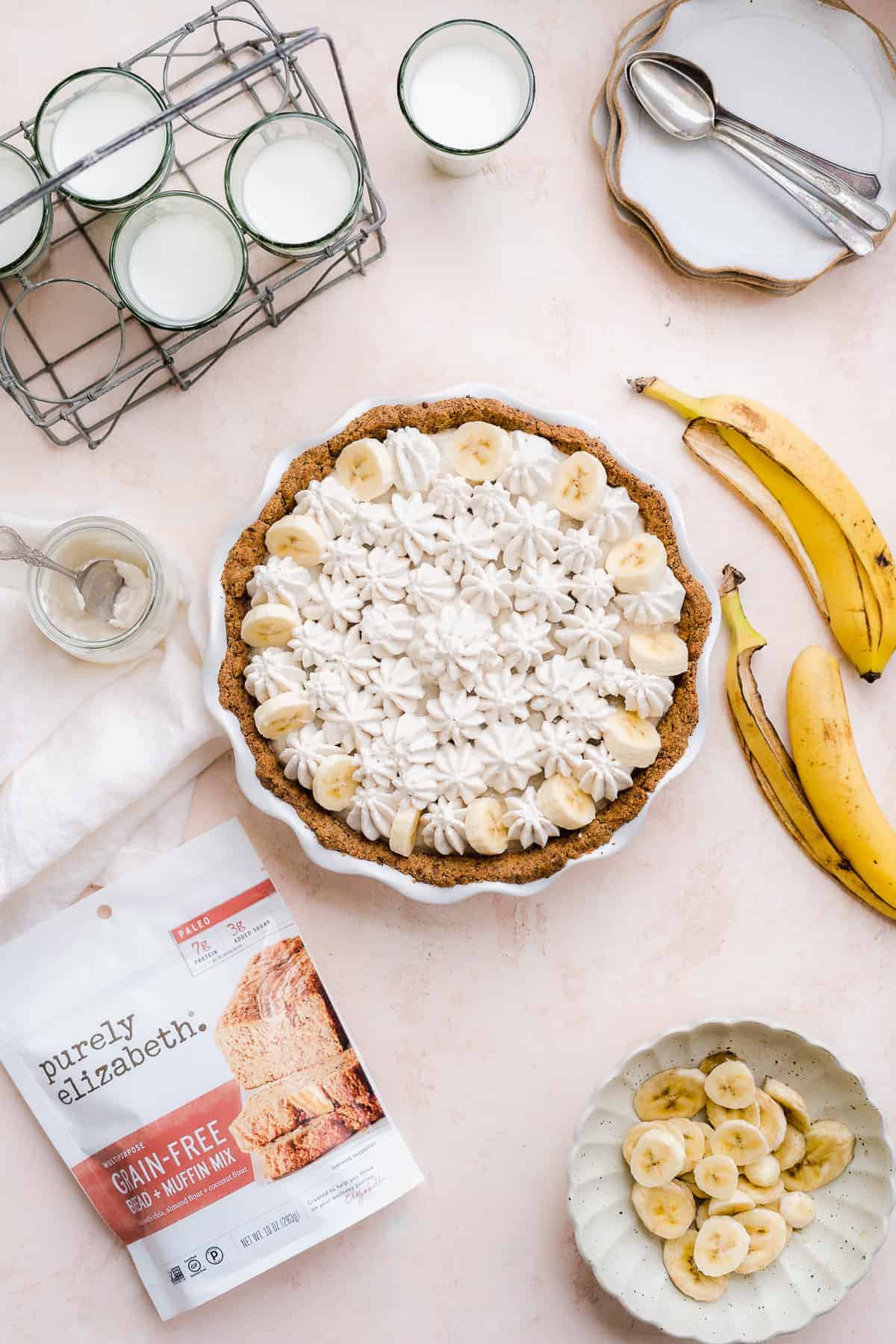 Banana pie on a pink surface with banana peels to the side and baking mix and plates.