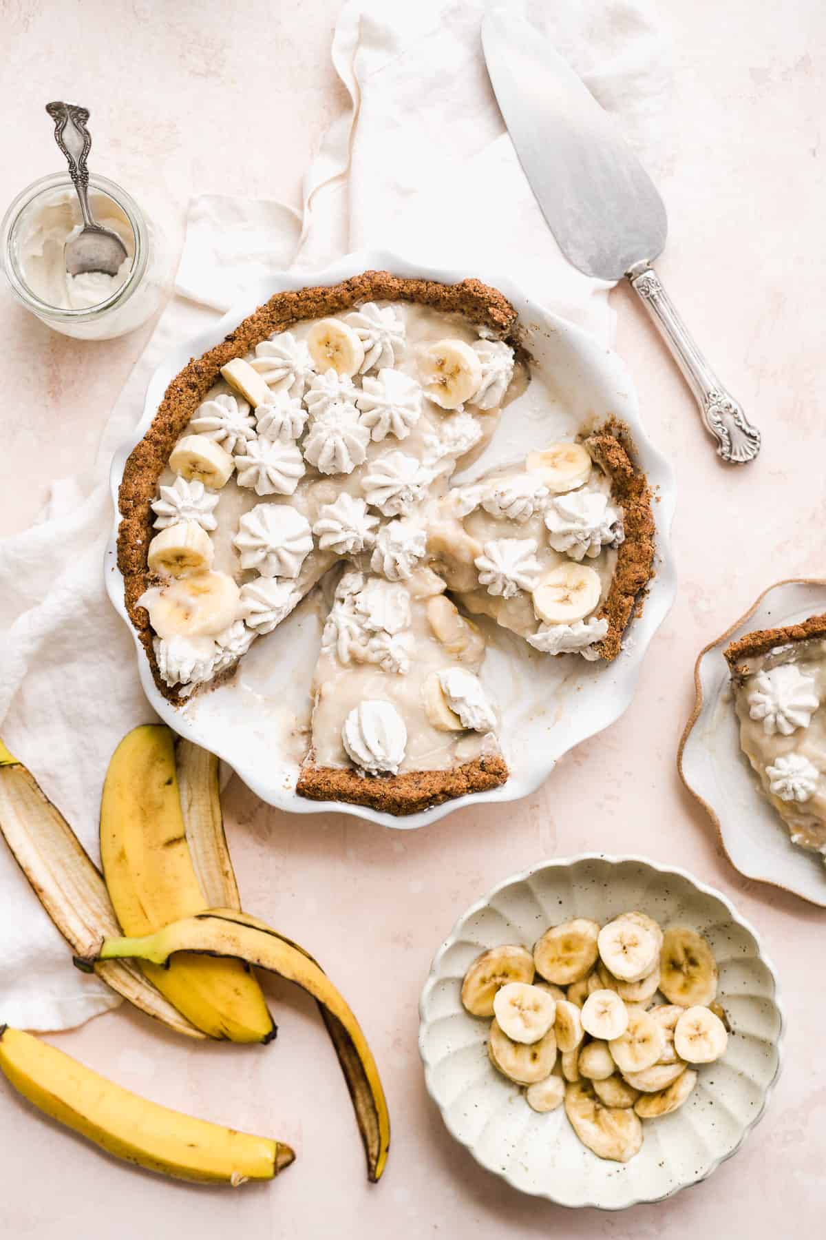 Banana pie sliced into pieces on a pink surface with banana slices and pie slices off to the side