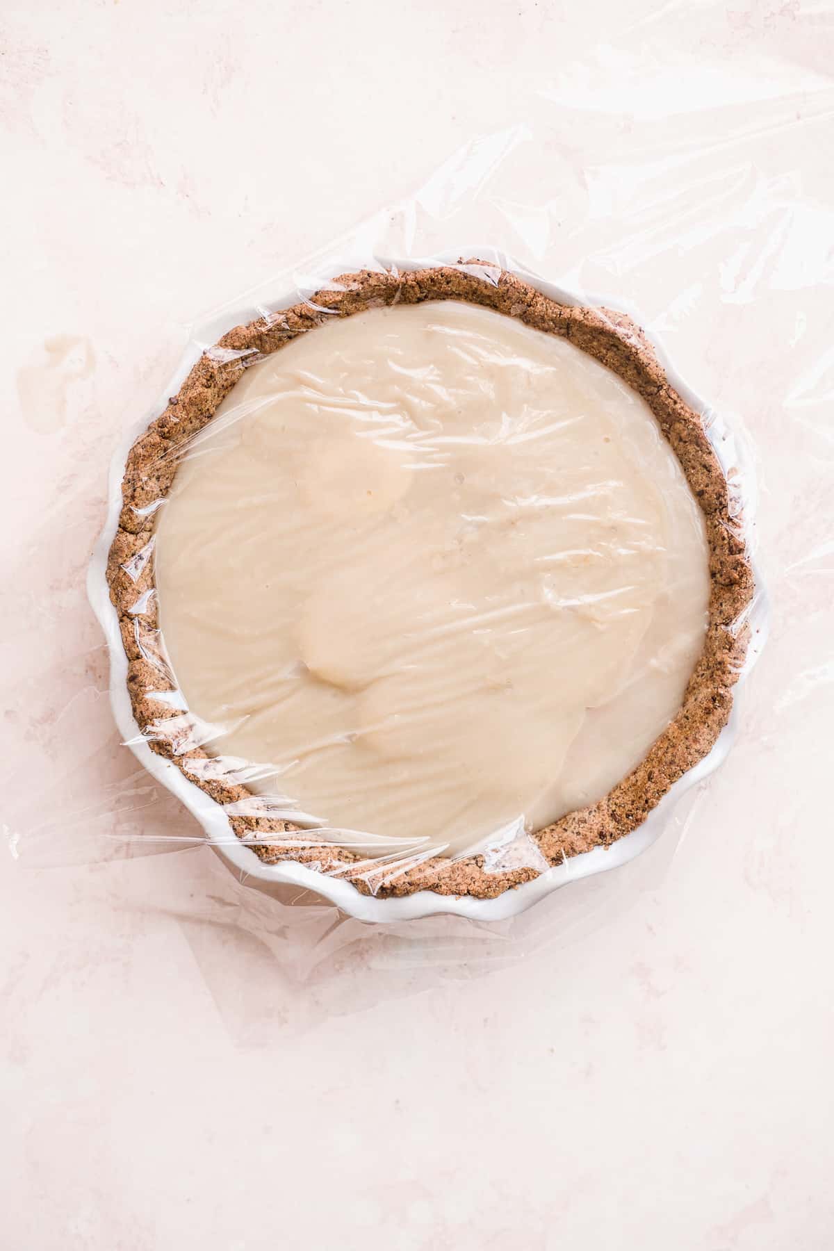 Image of pie on a pink surface with cling wrap over top of pie.