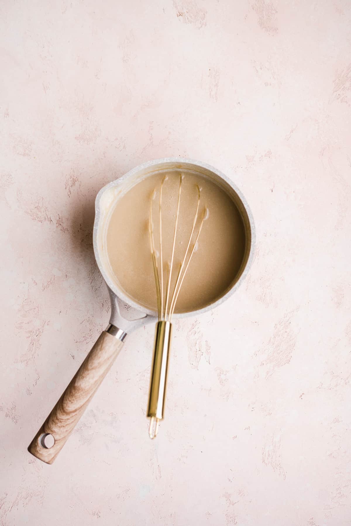 Pot with banana filing mixture with a gold whisk inside on a pink surface.