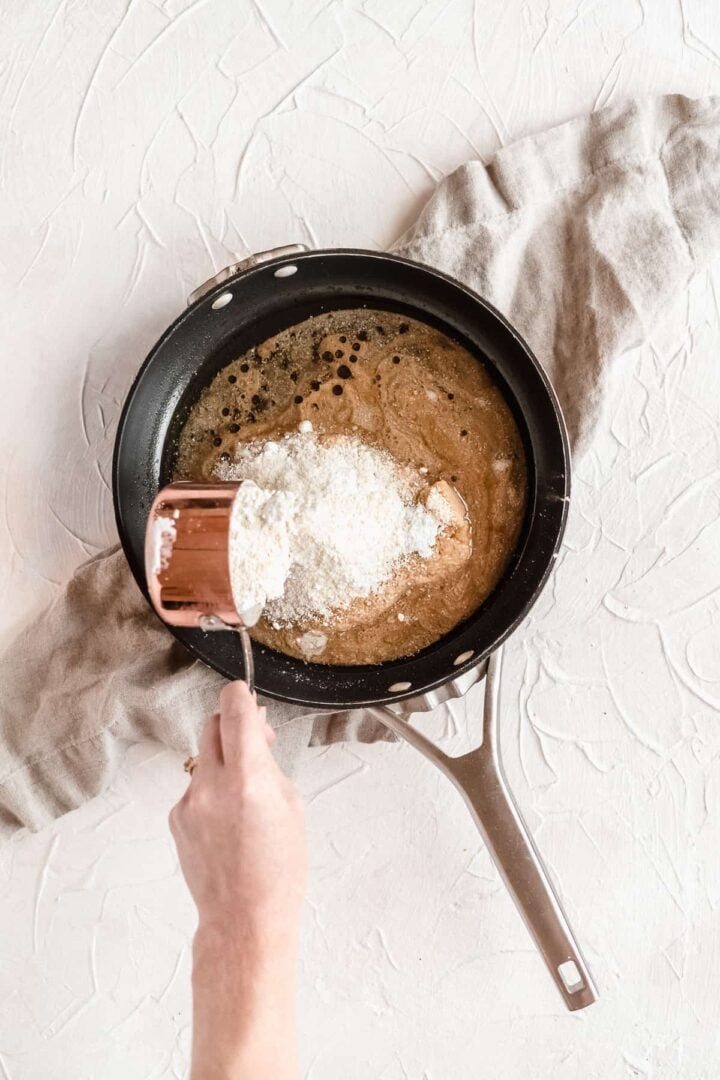 Hand pouring flour into a skillet with liquid.