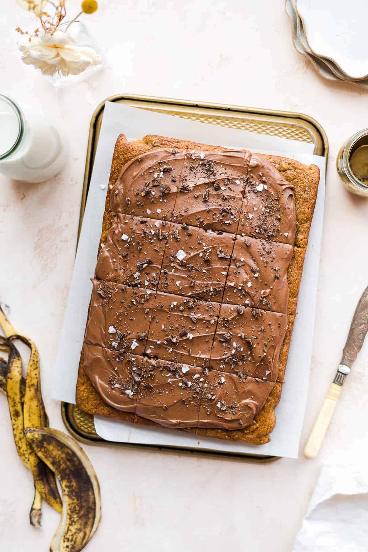 Banana cake with chocolate icing cut into squares on top of a pan with banana peel and milk off to the side.