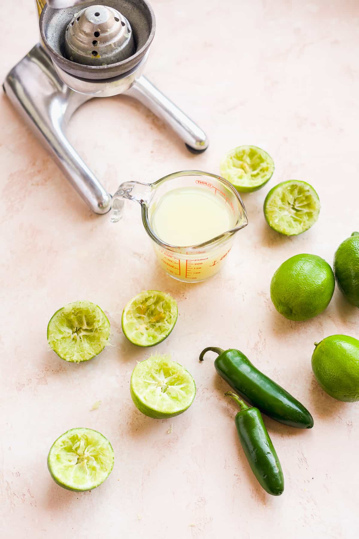 Measuring cup filled with lime juice in front of a metal juicer with limes on the surface.