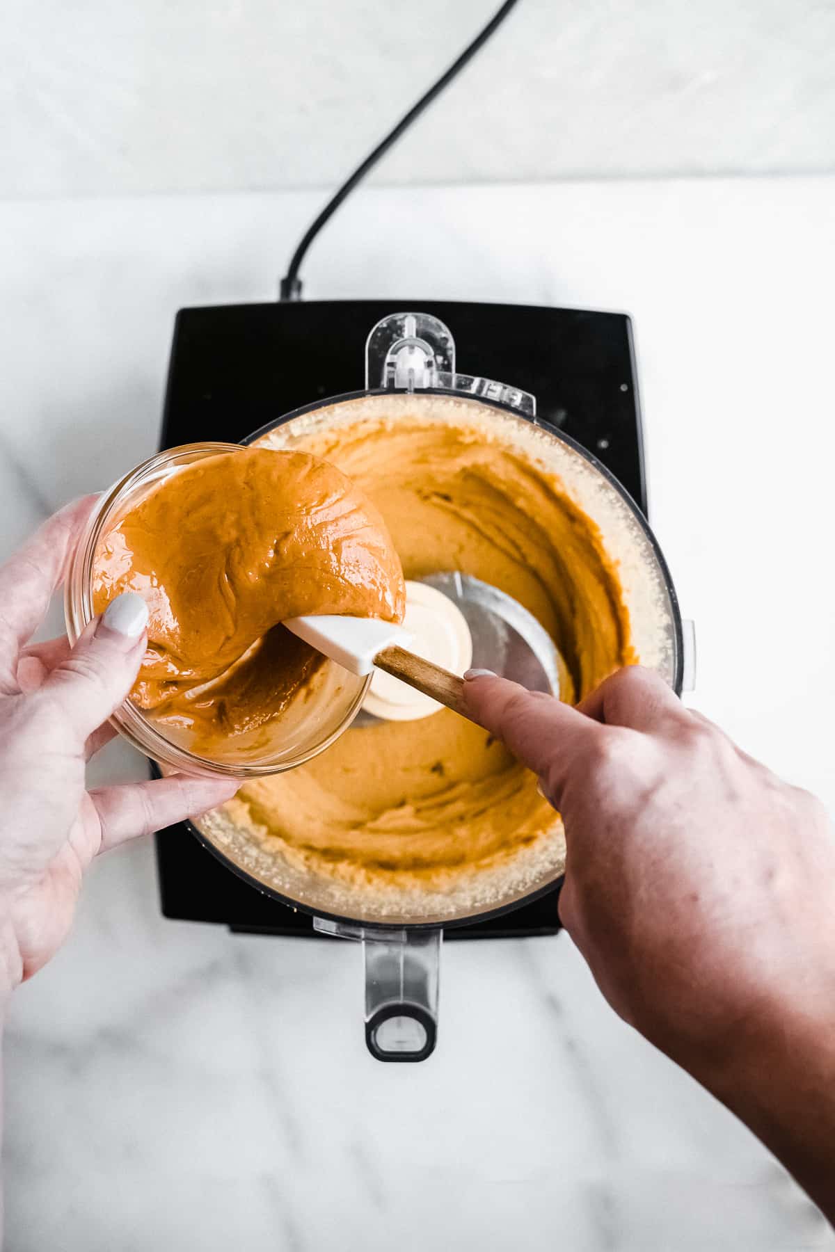 Hand pouring nut butter into food processor with tan mixture inside.