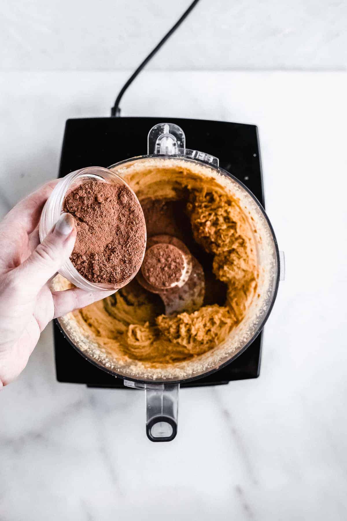 Hand pouring chocolate powder inside food processor with peanut butter mixture.