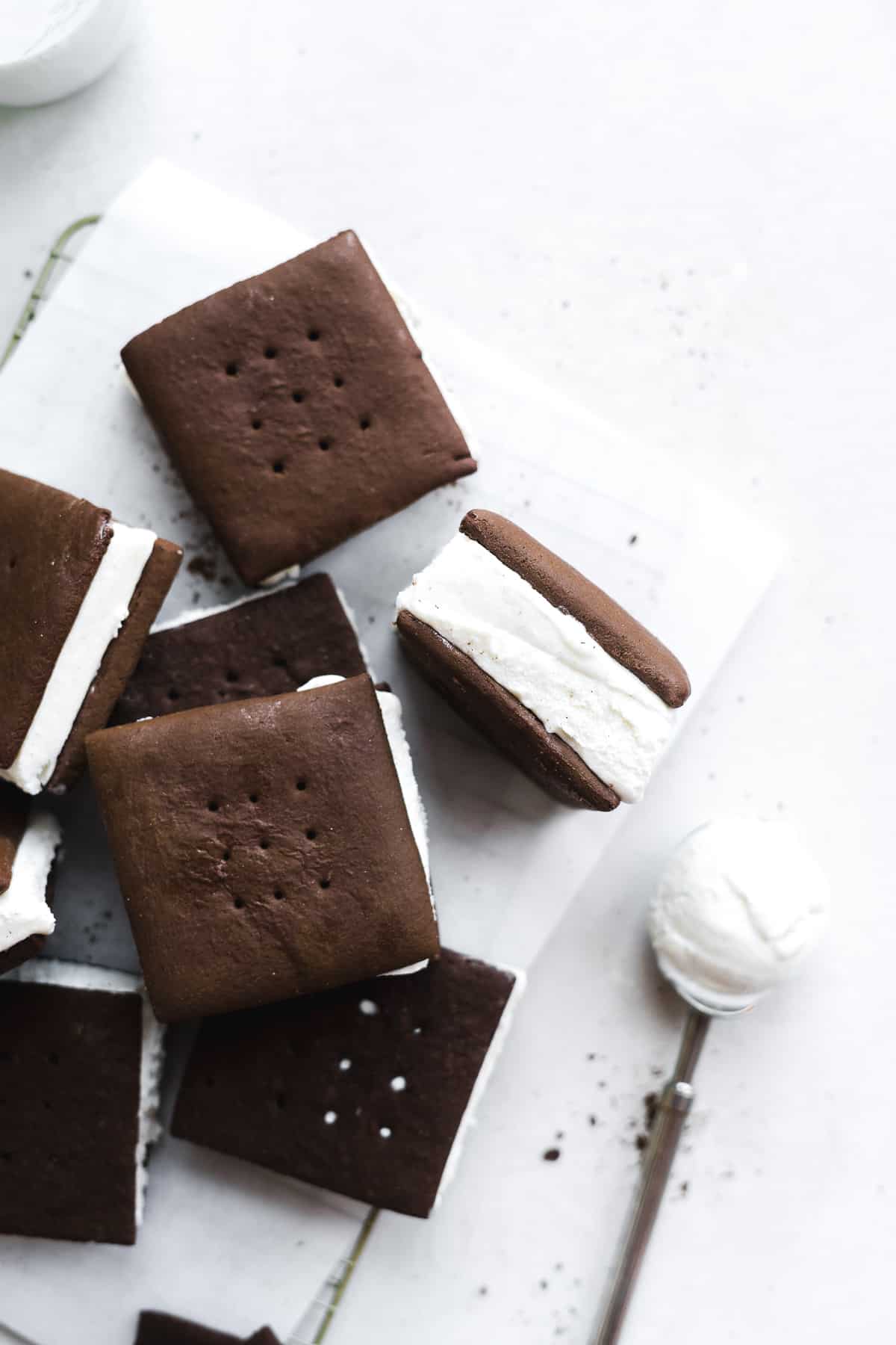 Chocolate ice cream sandwiches scattered on surface with one tipped upwards and an ice cream scoop on the side.