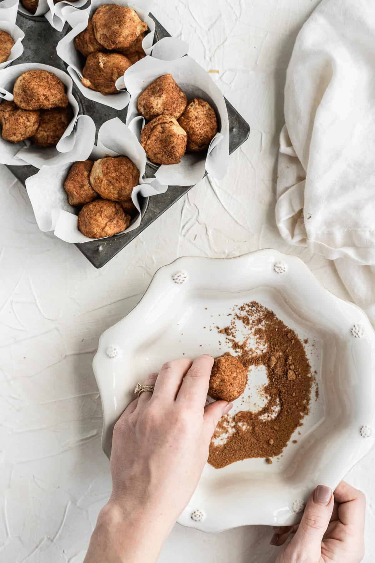 Hand rolling ball of dough in cinnamon with a muffin tin of to the side.