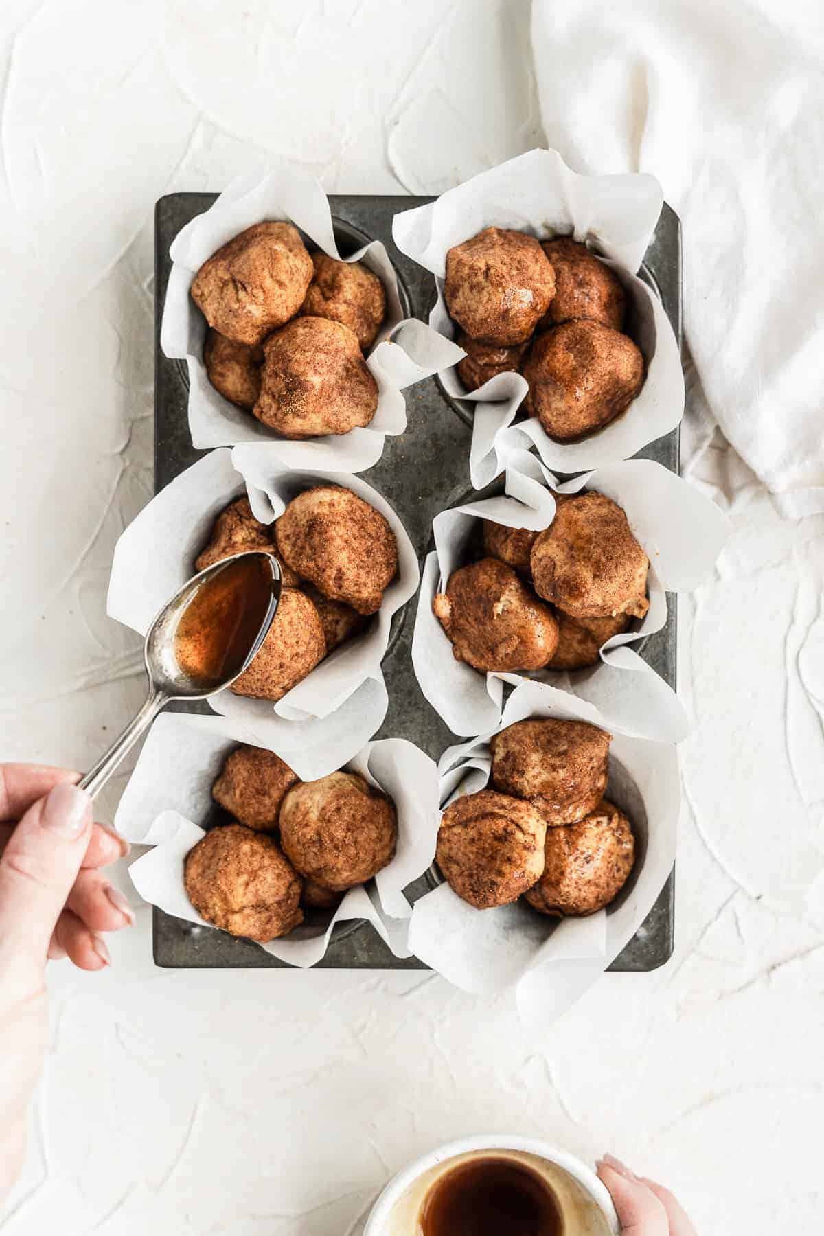 Hand drizzling maple syrup over muffins in a pan with white paper liners.