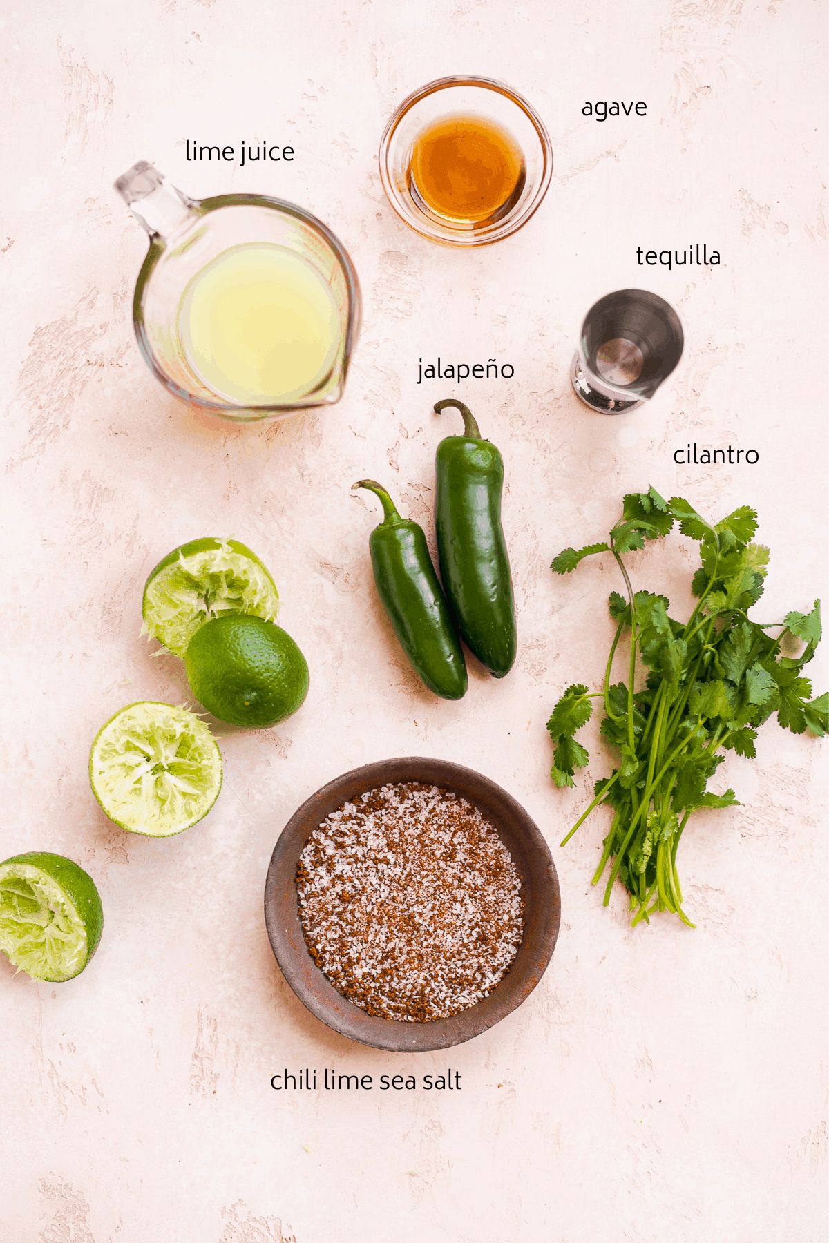Image of cocktail ingredients with black labels on a dusty pink surface including limes, cilantro, and jalapeño.