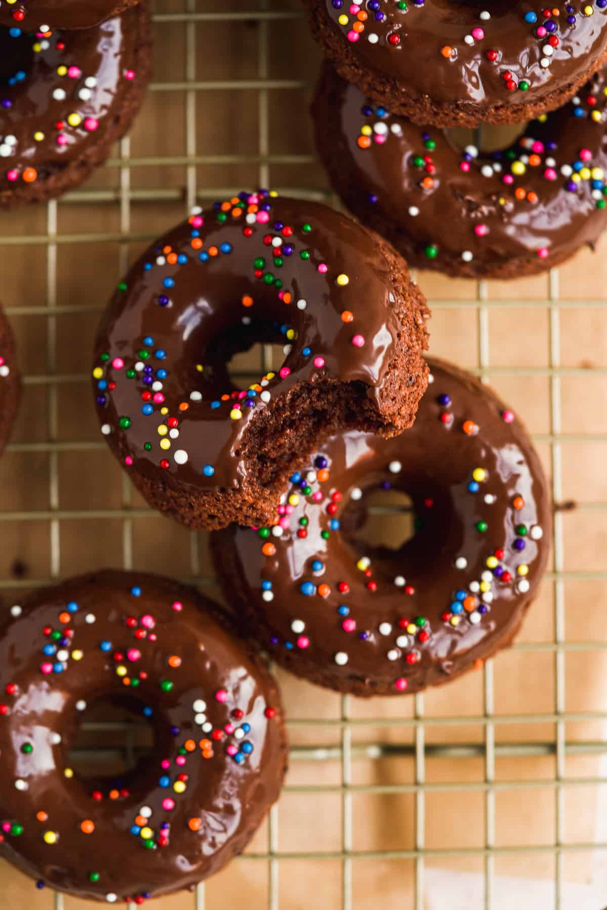 Chocolate doughnuts with sprinkles on wire rack with bite taken out of one.
