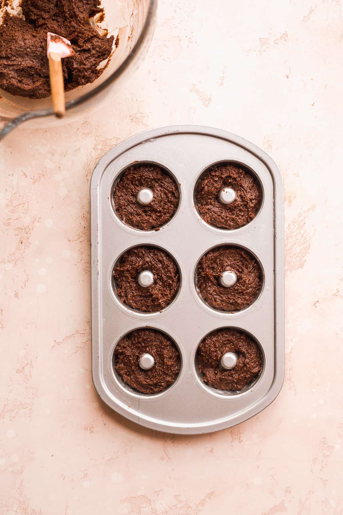 Doughnut pan filled with chocolate batter with bowl off to the side,