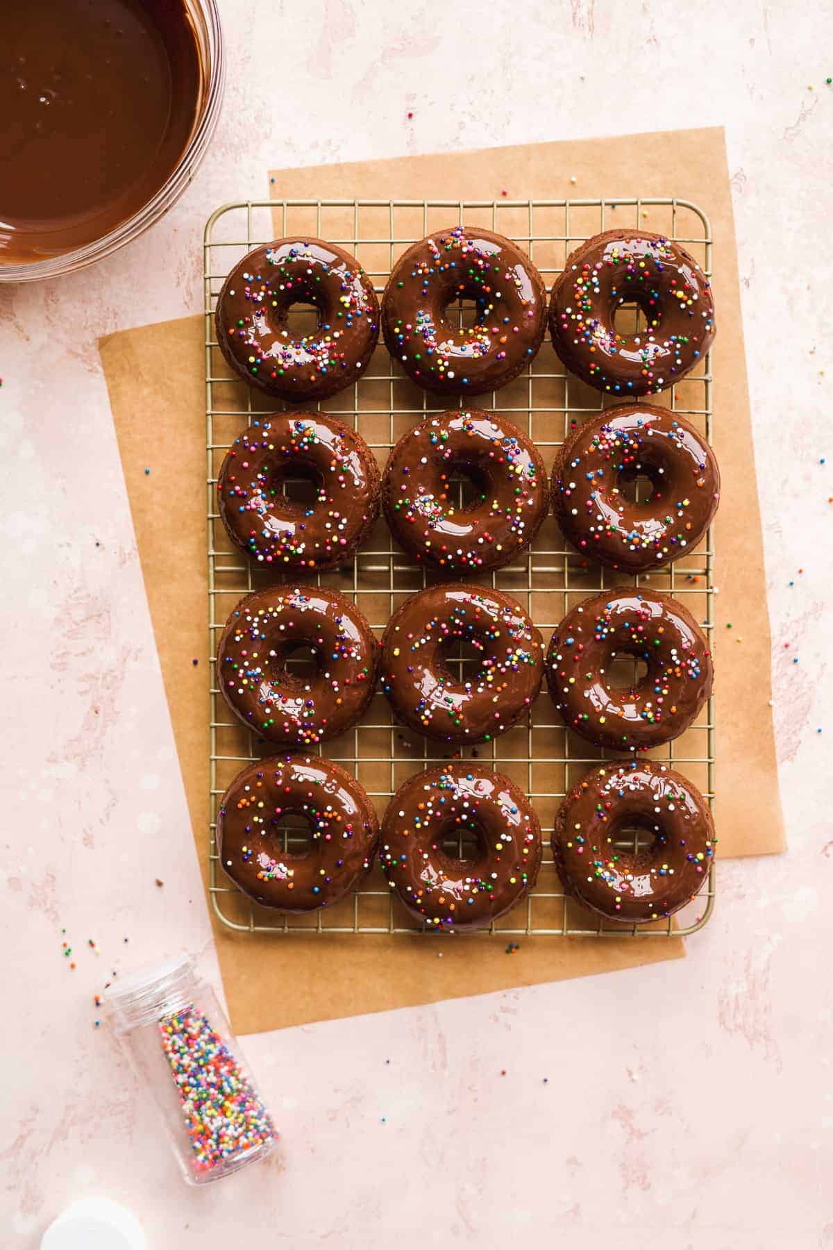 Chocolate doughnuts with sprinkles on wire rack with brown paper and chocolate in backgorund.