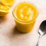 Glass jar of lemon curd with a spoon on the ground.