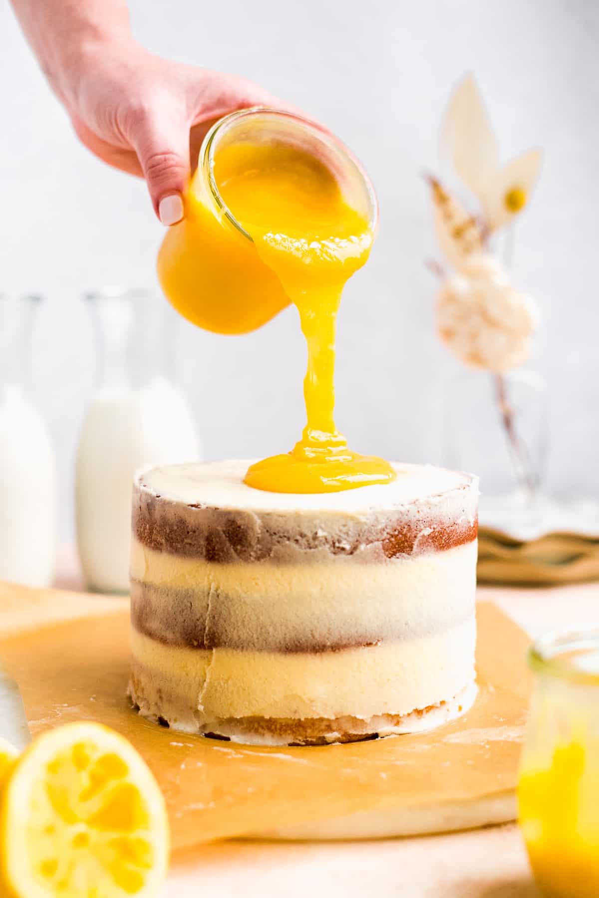 Hand pouring lemon curd over iced cake with milk and flowers in background.