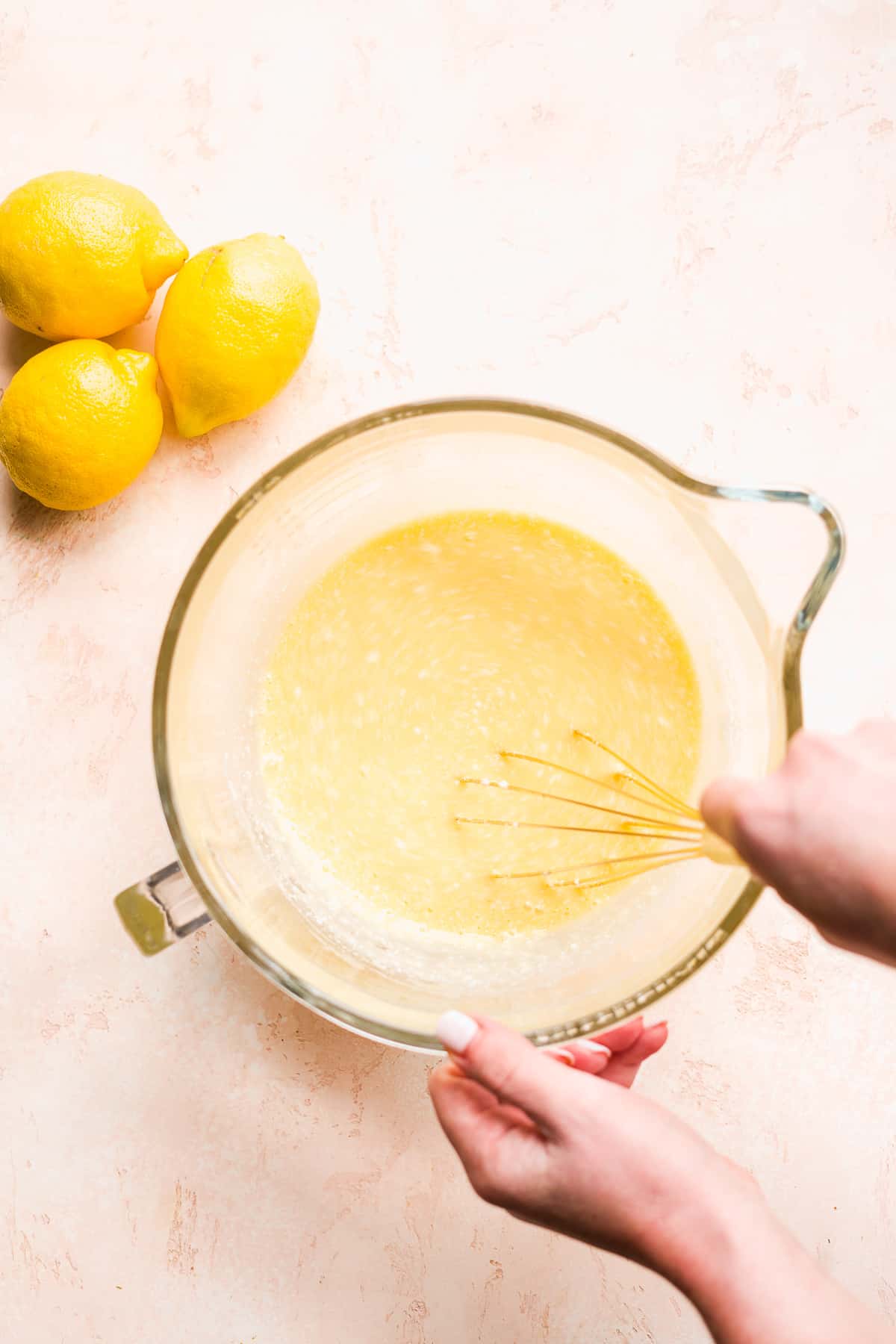 Hand whisking ingredients in a glass bowl with lemons on the side.