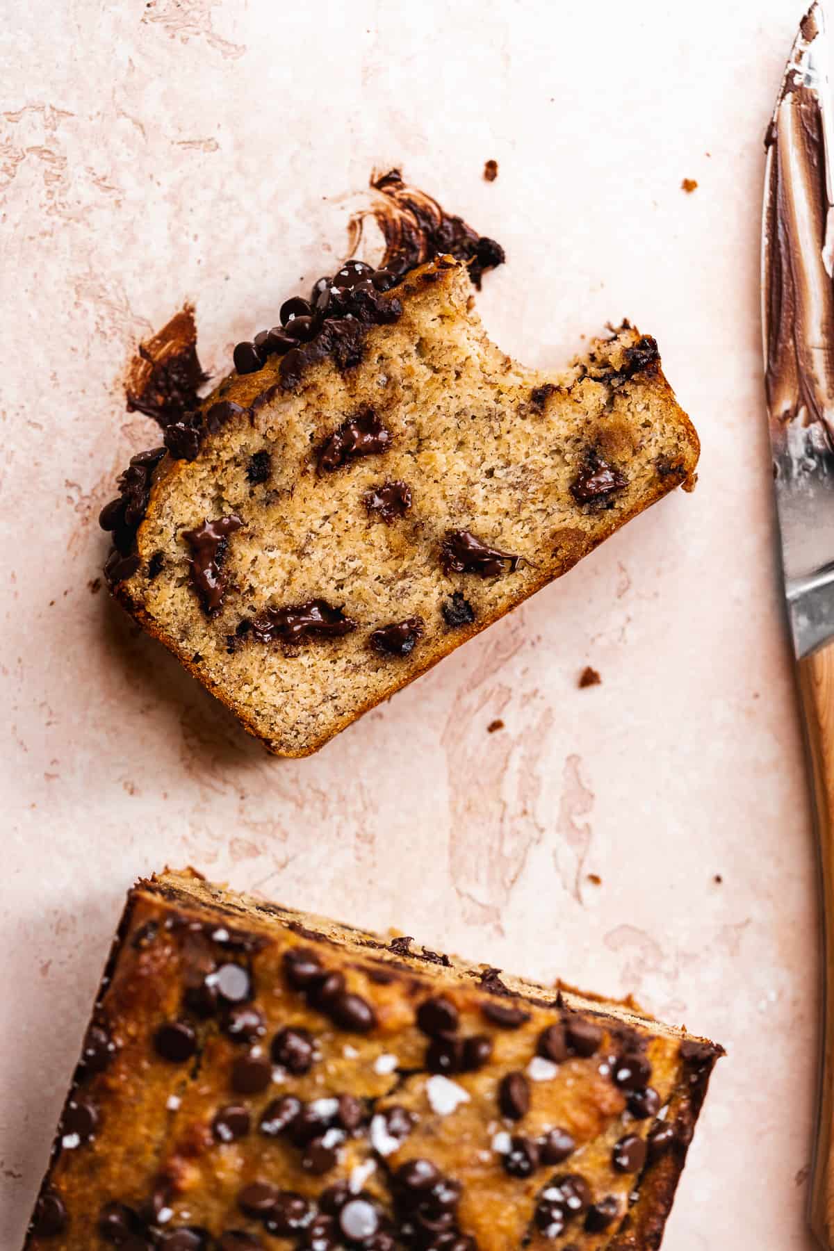 Slice of chocolate chip bread with a bite taken out of it on a pink surface with knife to the side.