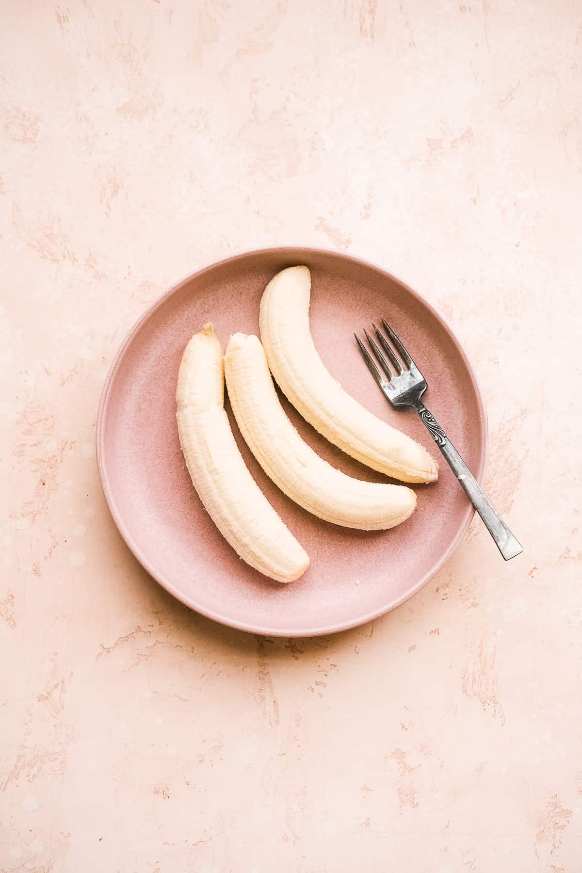 Three bananas on a pink plate with a fork on the side.
